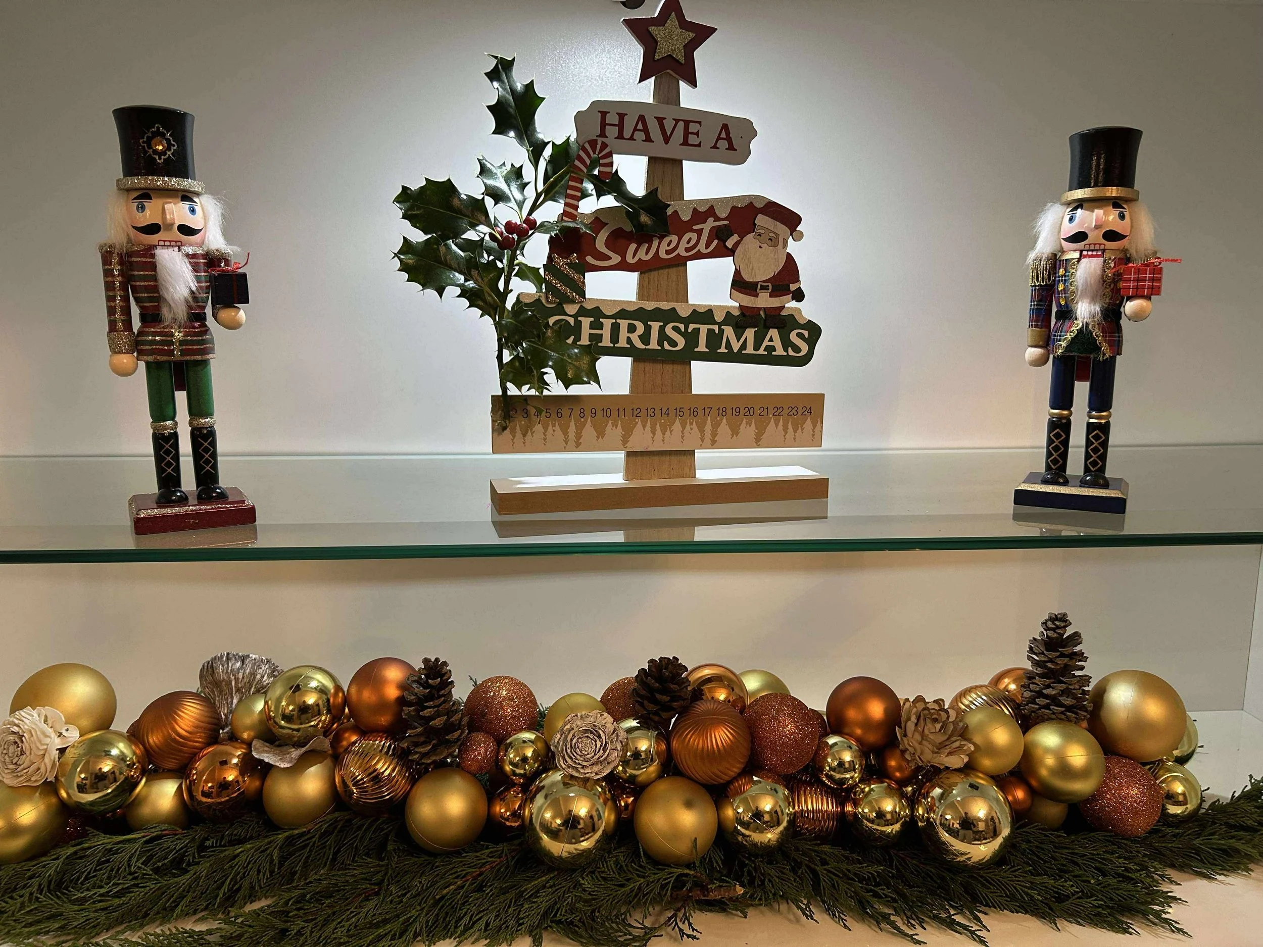 Décoration de Noël avec deux soldats en bois, un panneau de Noël avec un message festif, des branches de houx, et une guirlande de boules de Noël dorées, brunes et or iridescentes