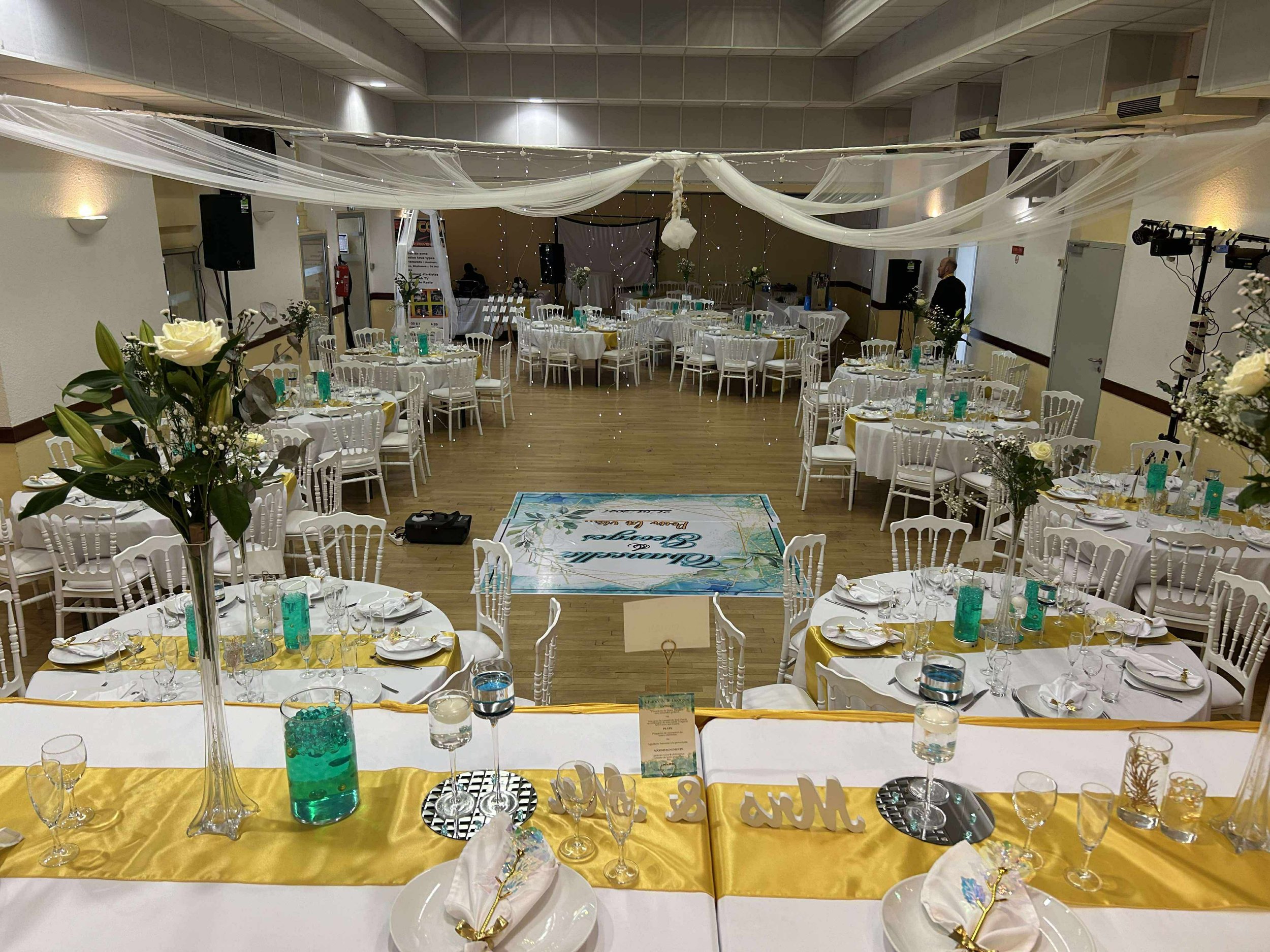 Salle de réception décorée pour un mariage, avec des tables rondes ornées de fleurs blanches, verres, assiettes et couverts, suspension de tulle blanc au plafond, Clés de table avec le mot 'Love', et un espace central vide pour la danse.