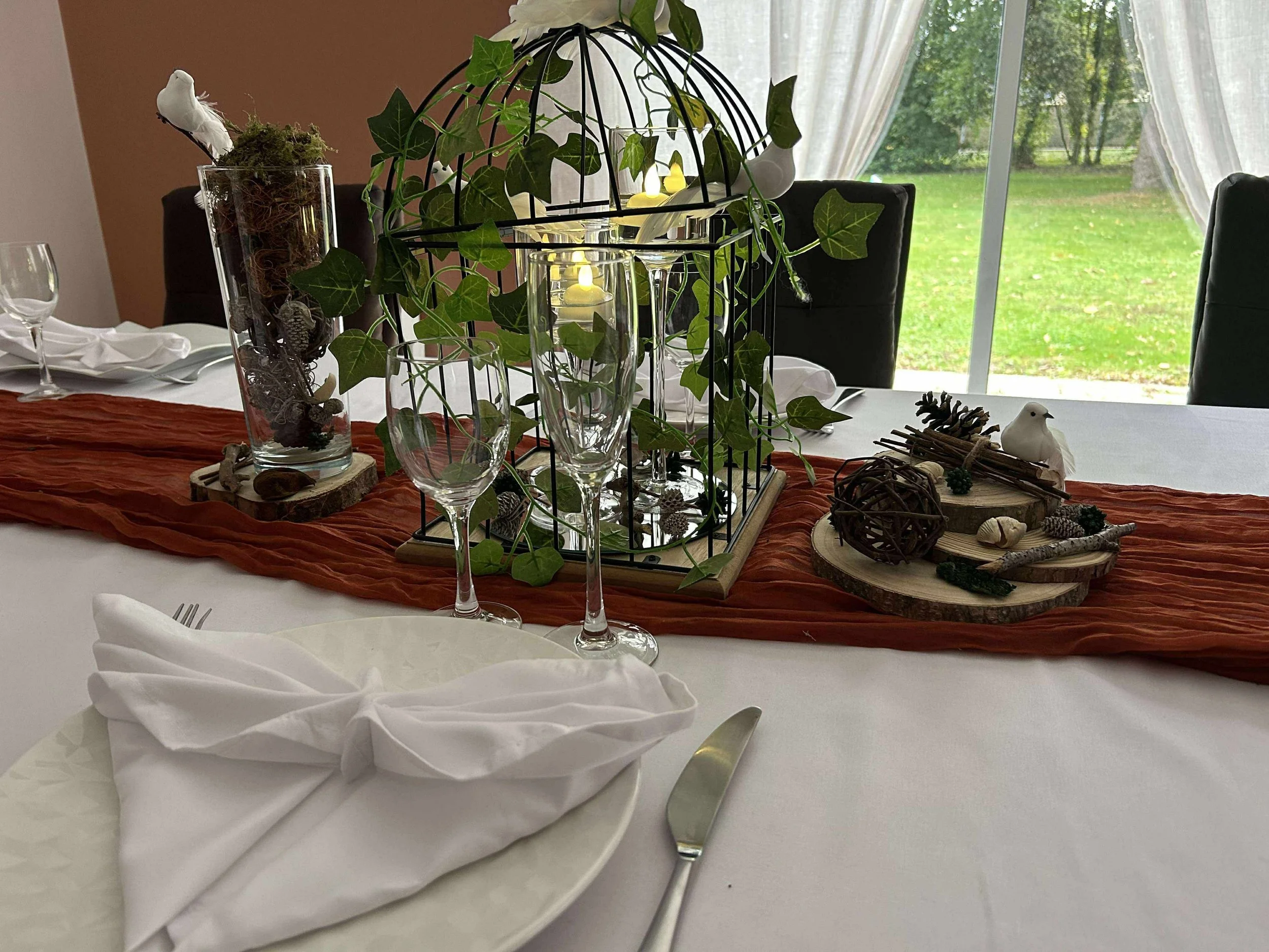 Une table de fête décoration rustique avec une chemin de table en tissu orange, des éléments naturels, des branches, et un centre de table avec des plantes vertes, des bougies et des éléments en bois. La pièce a de grandes fenêtres avec des rideaux b