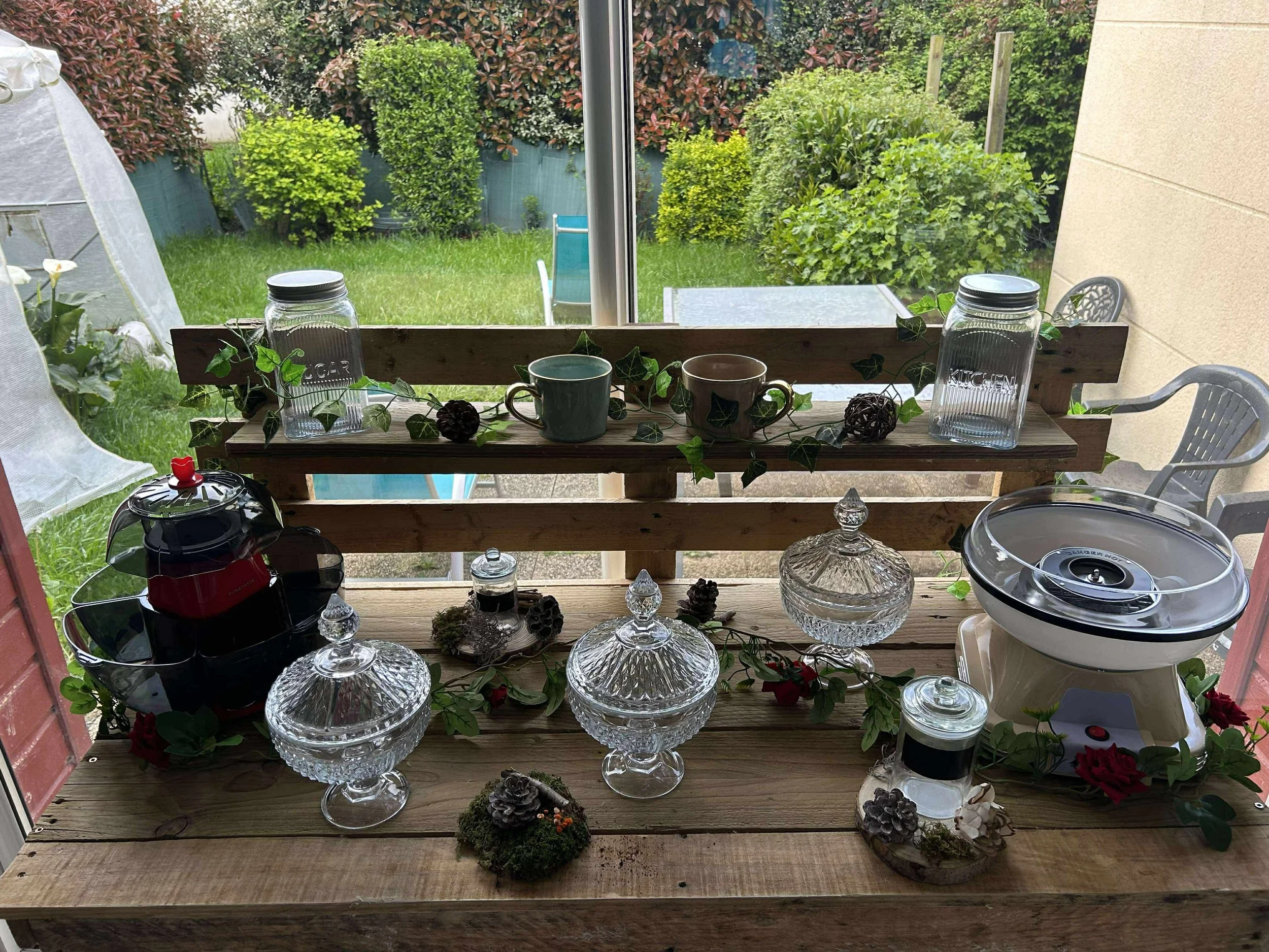 Table en bois avec des verreries, tasses, et décorations florales, devant une fenêtre donnant sur un jardin verdoyant.