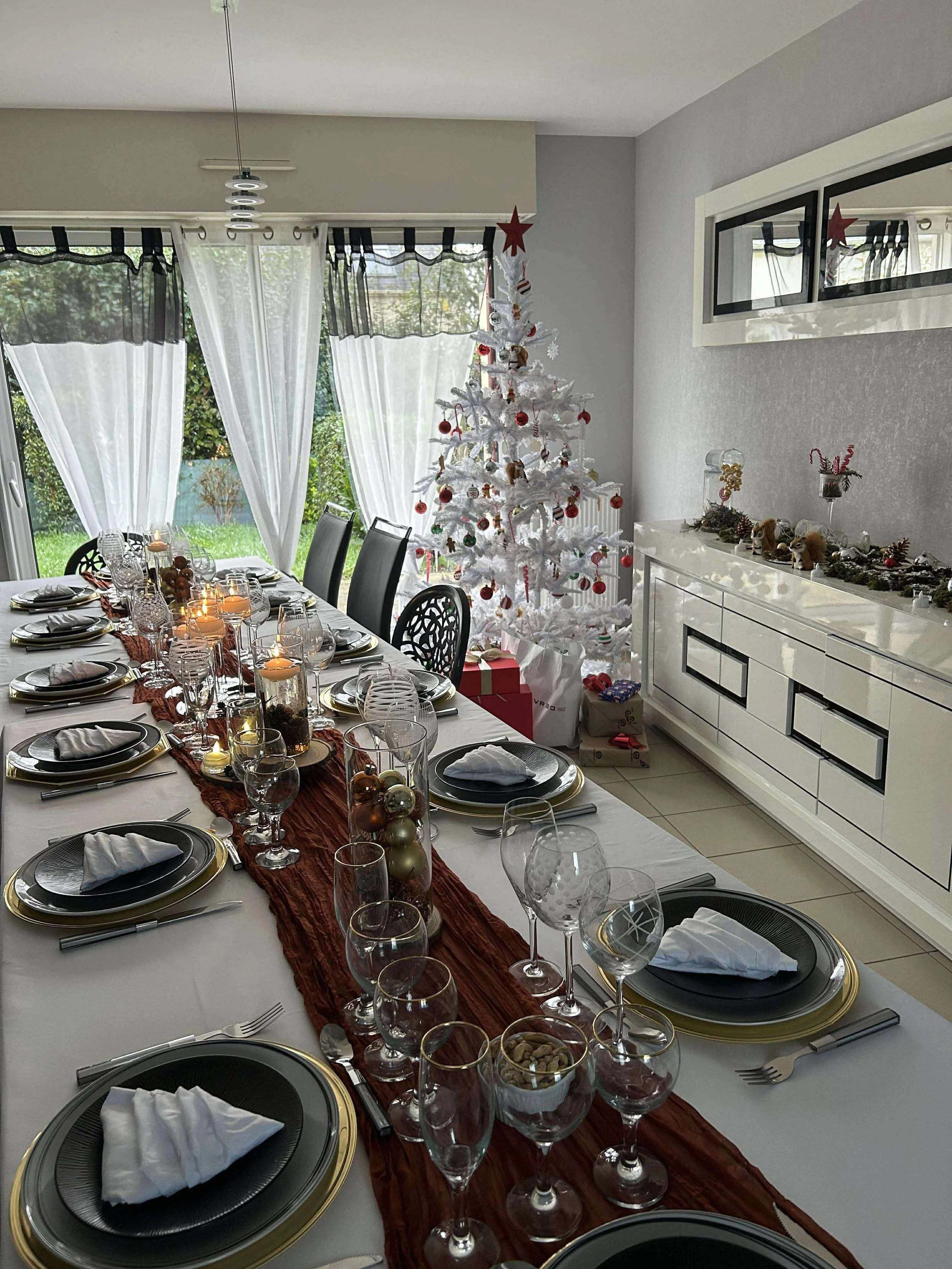 Salle à manger décorée pour Noël avec un sapin blanc orné de décorations rouges et or, une table festive avec des assiettes, des verres et des bougies, et une cheminée décorée avec des objets de Noël.