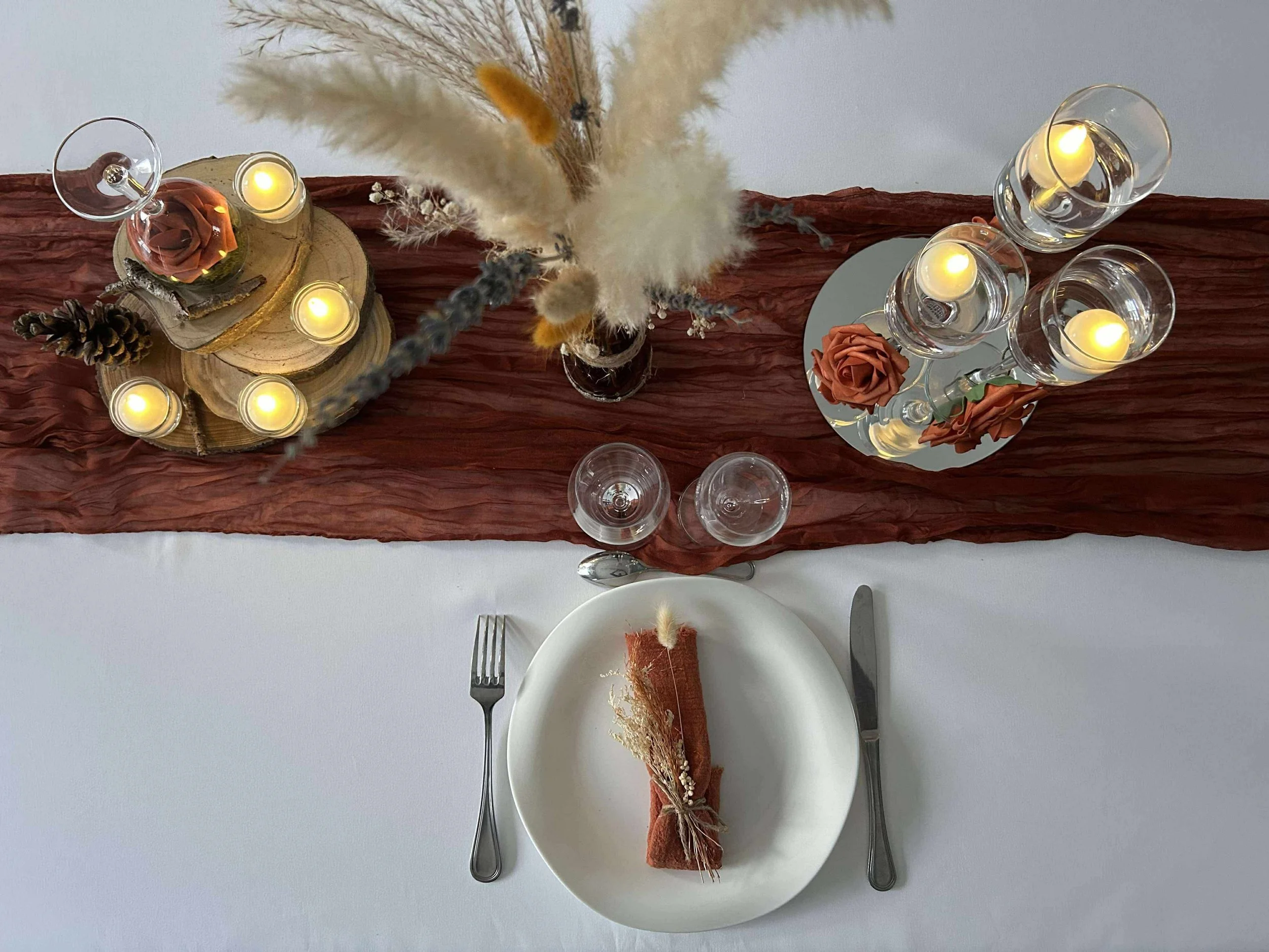 Table de fête avec une nappe blanche, un centre de table avec des bougies, des fleurs et des décorations en bois, un verre à vin, des verre à eau, une assiette avec un nappage enroulé, et des couverts.