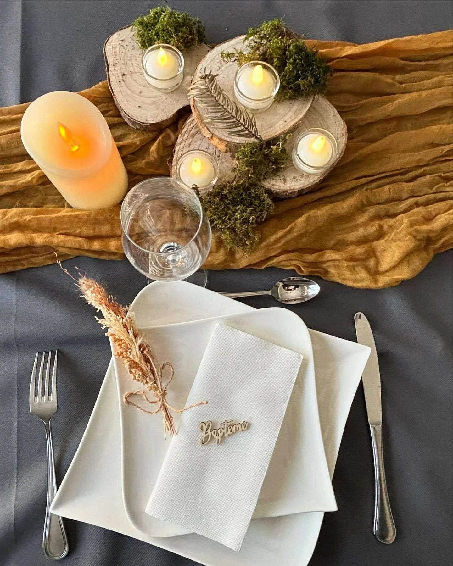 Table de fête avec un décor de bougies, serviettes et menu, ornée de tissus dorés, de bois, de mousse et de plantes vertes.