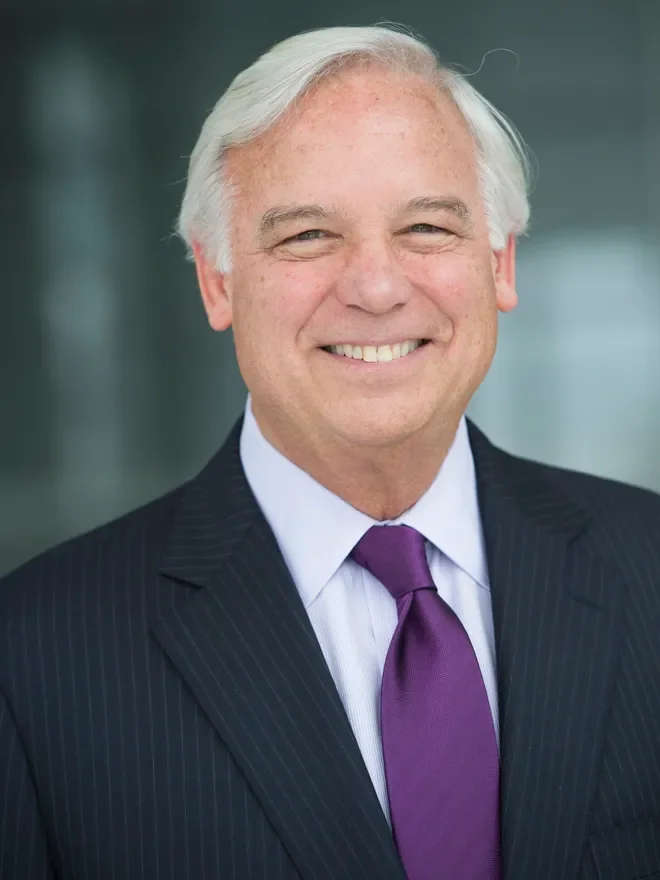Smiling older man with white hair wearing a dark pinstripe suit, a white shirt, and a purple tie, standing outdoors.