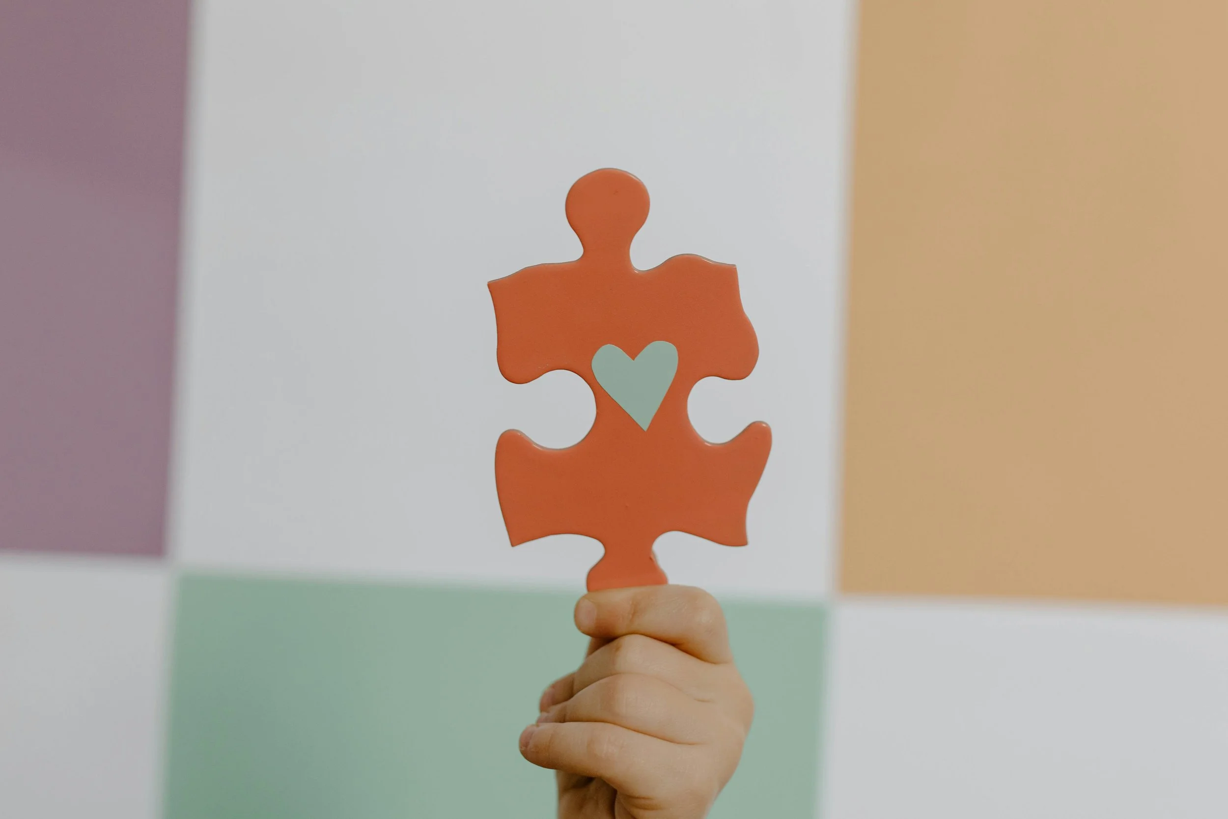 A hand holding a peach-colored puzzle piece with a light blue heart in the center against a colorful geometric background.