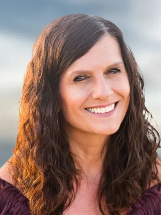 A woman with long wavy brown hair and a bright smile outdoors during sunset, wearing a dark top.