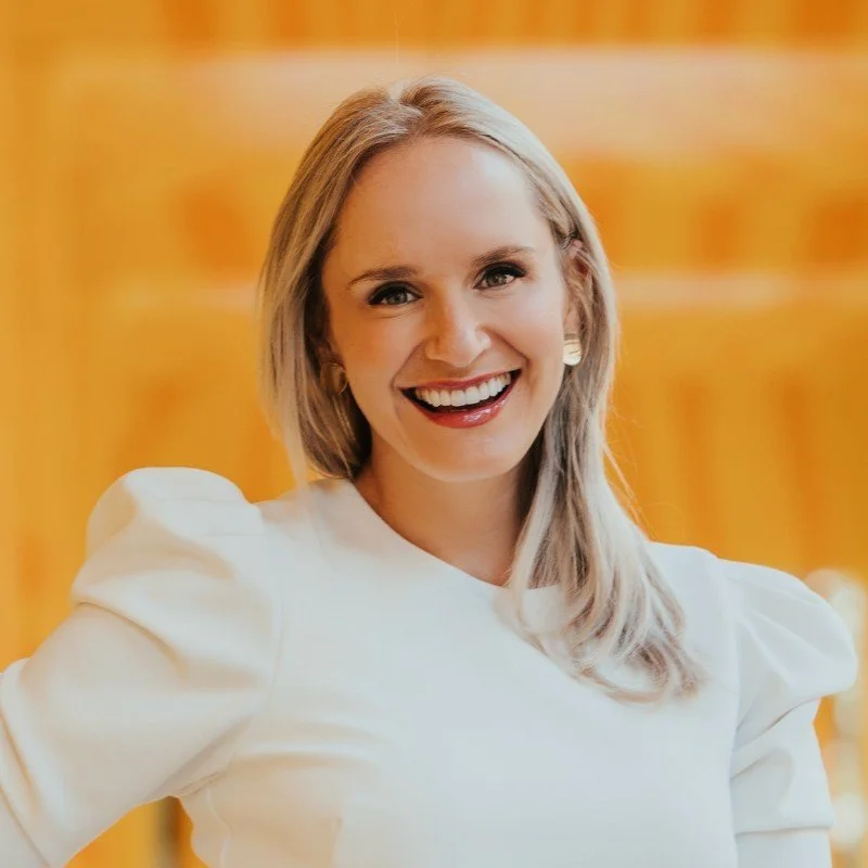 A woman smiling with blonde hair and wearing a white top standing in front of a warm, blurred background.