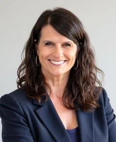 A woman with shoulder-length dark brown hair, wearing a navy blazer and a blue top, smiling against a light background.