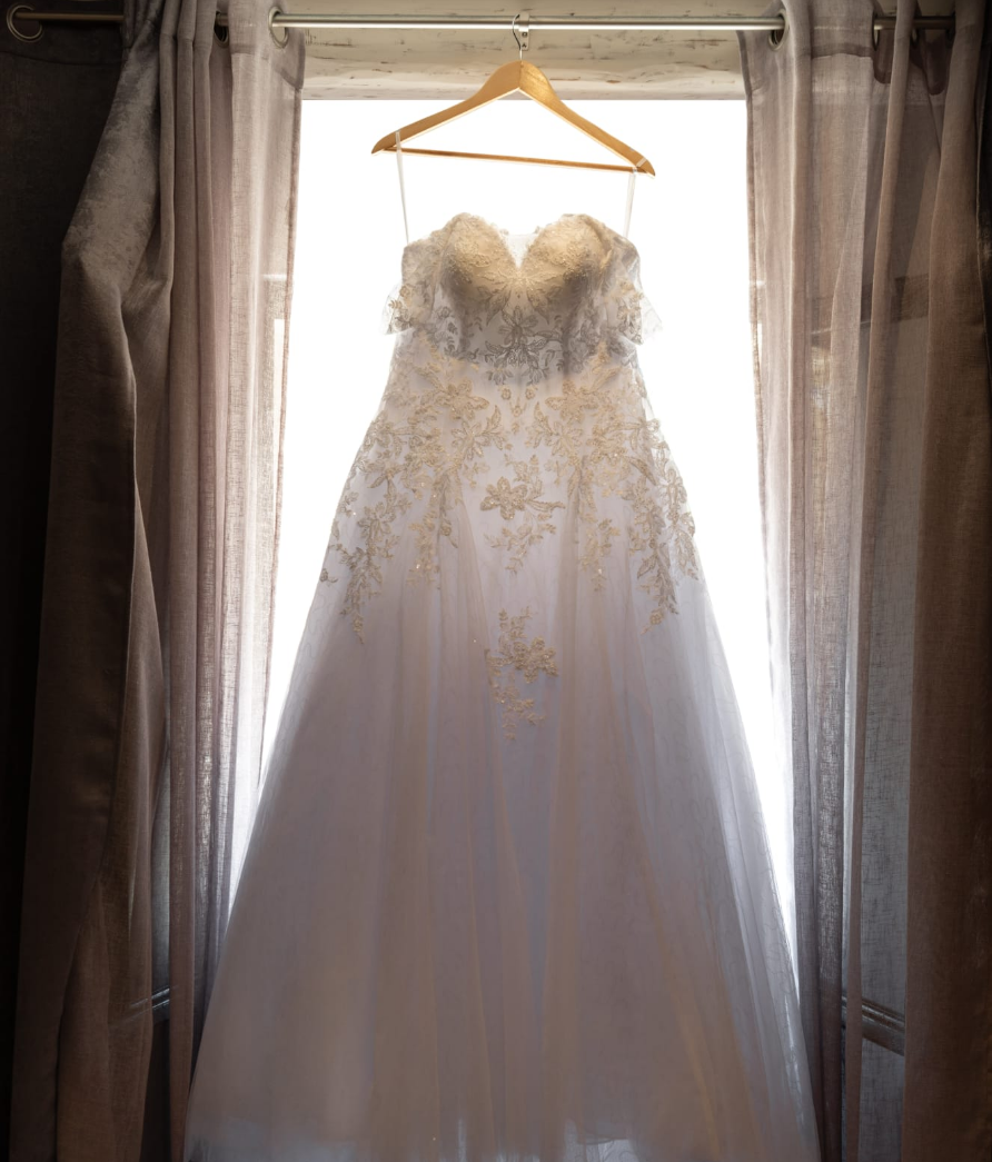 Robe de mariée longue bustier en dentelle, suspendue sur un cintre en bois à la fenêtre de la chambre nuptiale au château de Roquefeuille, mariage organisé par A&L Event.