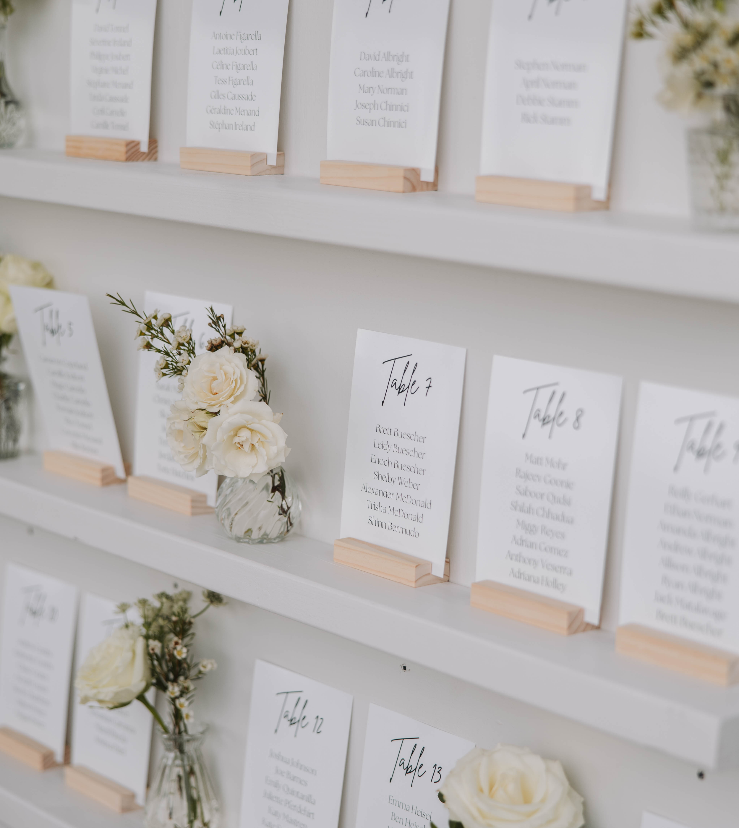Plan de table en bois avec étagères décorées de panneaux indiquant les numéros de table et les noms des invités, agrémenté de petits vases fleuris aux tons vert d’eau, blanc et eucalyptus, réalisé par A&L Events pour un mariage élégant.
