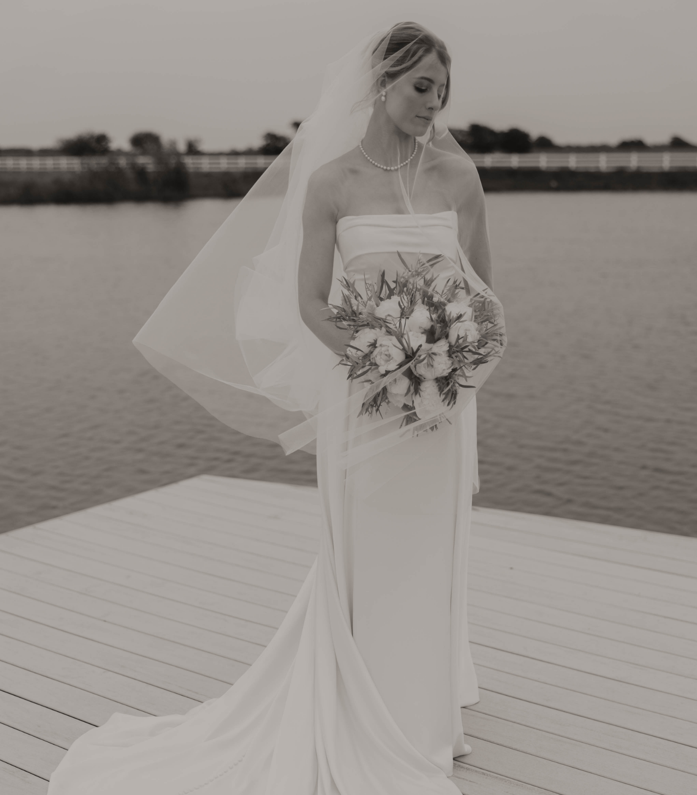 Une mariée en robe blanche bustier avec un voile, tenant un bouquet de fleurs, devant un plan d'eau sur un ponton , en noir et blanc.