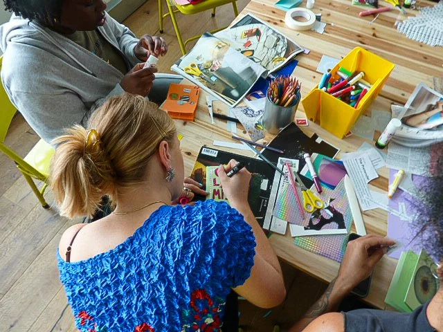 A birds eye image of an arts and crafts table with papers, magazines and pens on it. 3 people sit around the table, doing arts and crafts