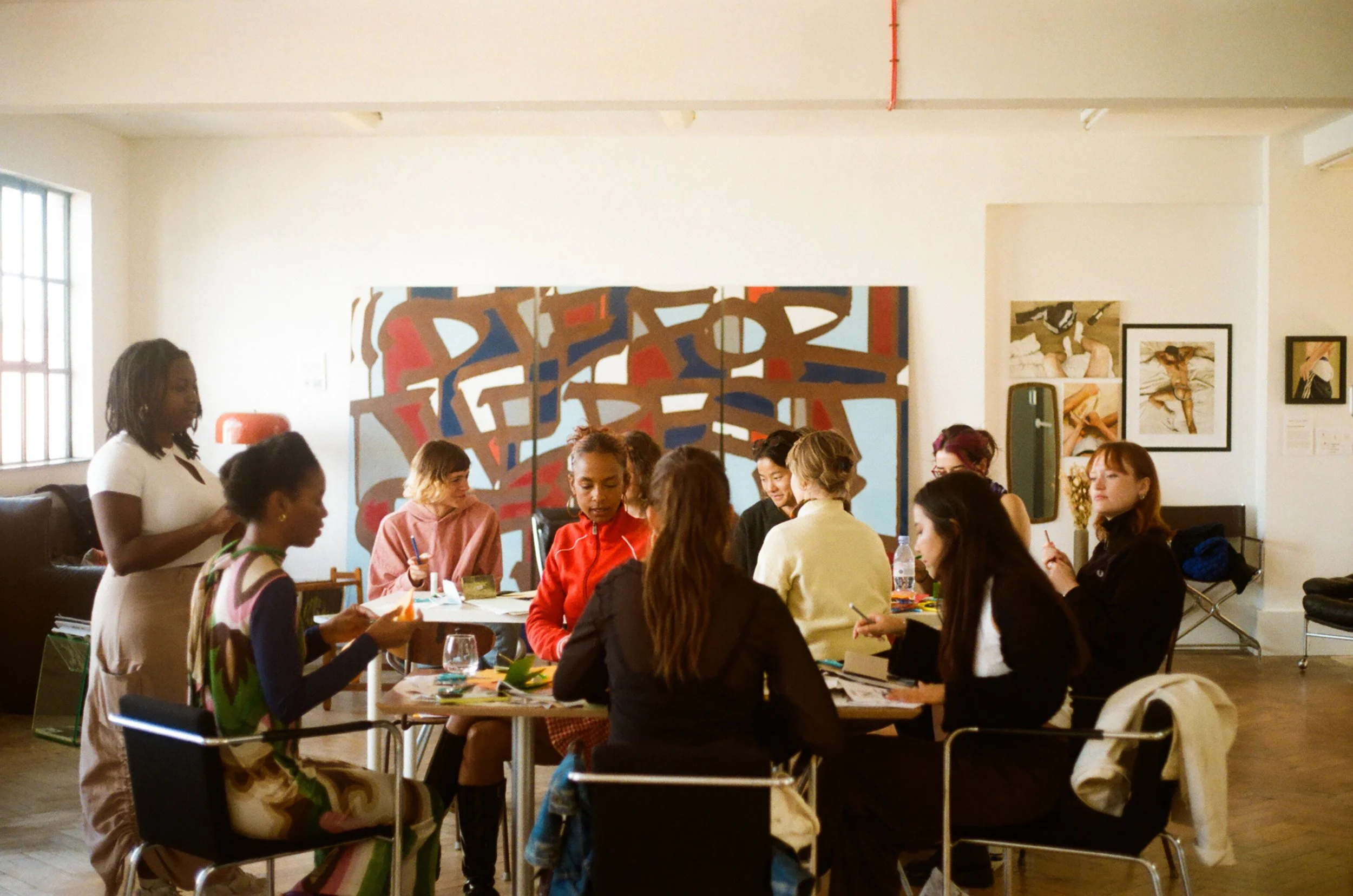 An image of a group of women sitting around a large table in a room with paintings on the walls. There's a woman standing at the head of the table, hosting the workshop.