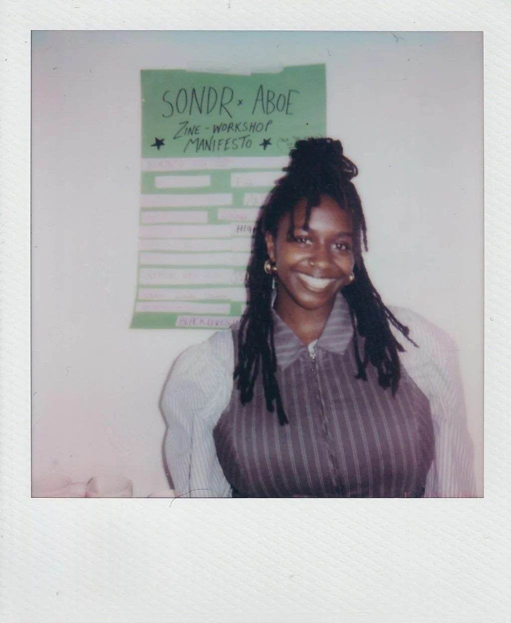 A polaroid image of a woman with a big smile, standing in front of a poster that says 'SONDR X ABOE' Zine Workshop Manifesto