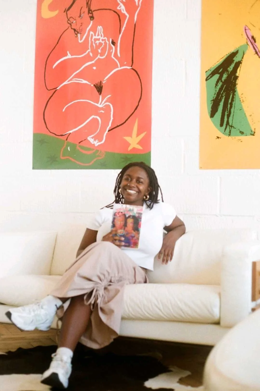 A woman sitting on a sofa smiling, with vibrant paintings on the wall behind her.