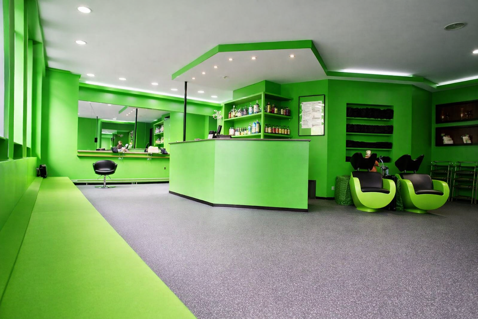 Ein Friseursalon mit grüner Wandfarbe, Empfangstresen, zwei schwarze Friseurstühle, eine schwarze Sitzbank, Spiegel, und eine Lith sowie einige Regale mit Produkten.