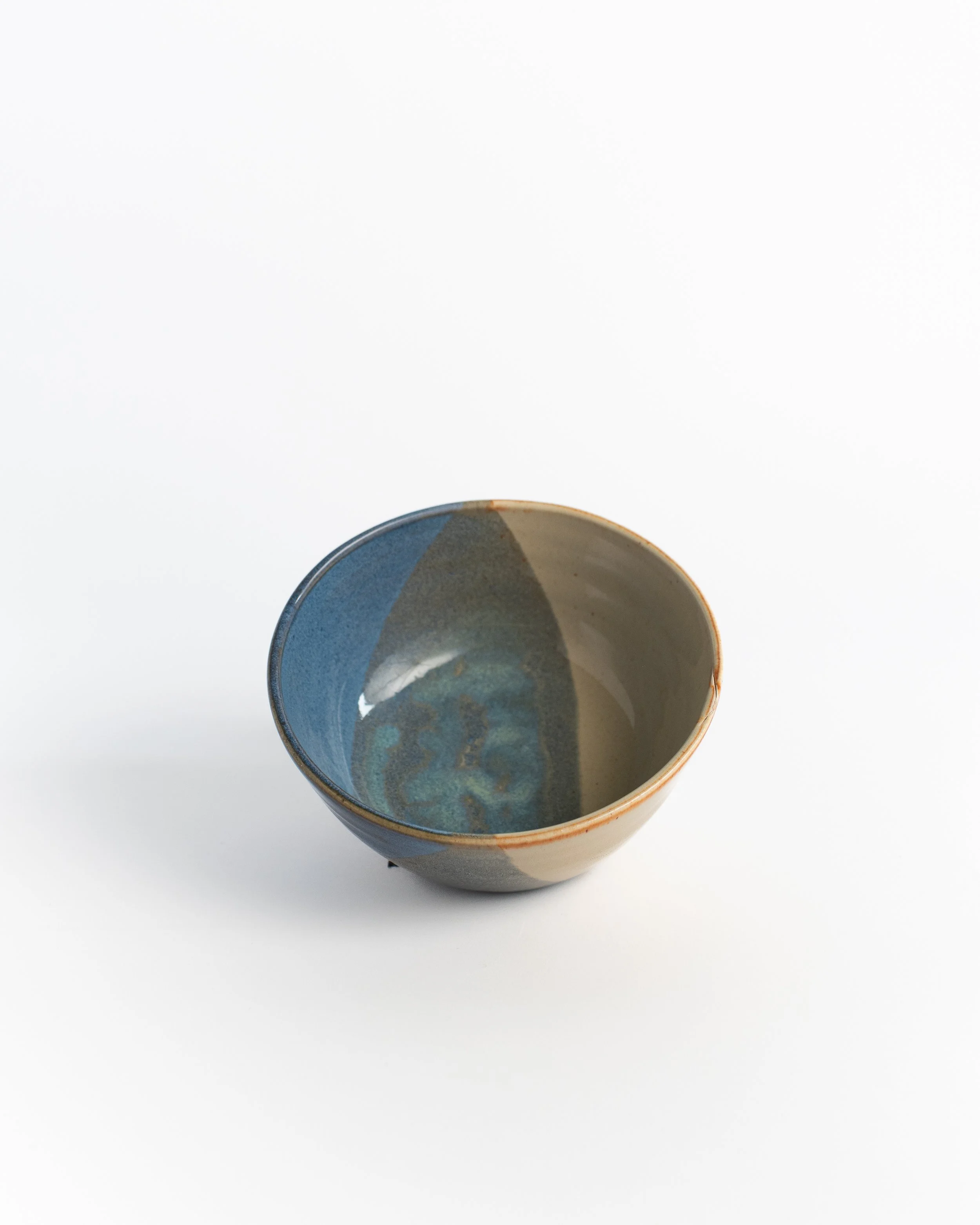 Rounded ceramic bowl seen from above, blue-grey and sand glaze meeting across the interior.