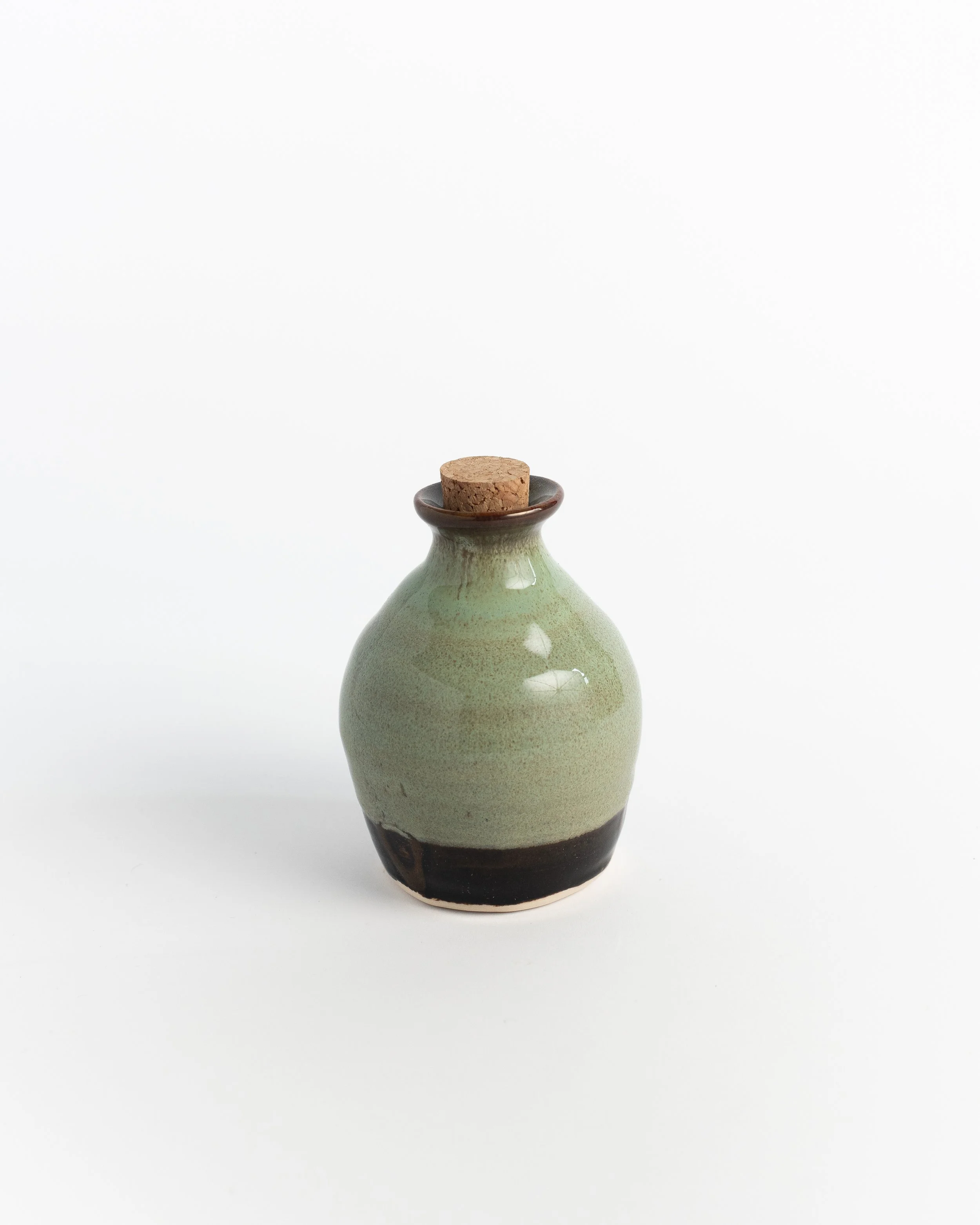 Rounded corked ceramic bottle in soft moss green over a dark chocolate foot.