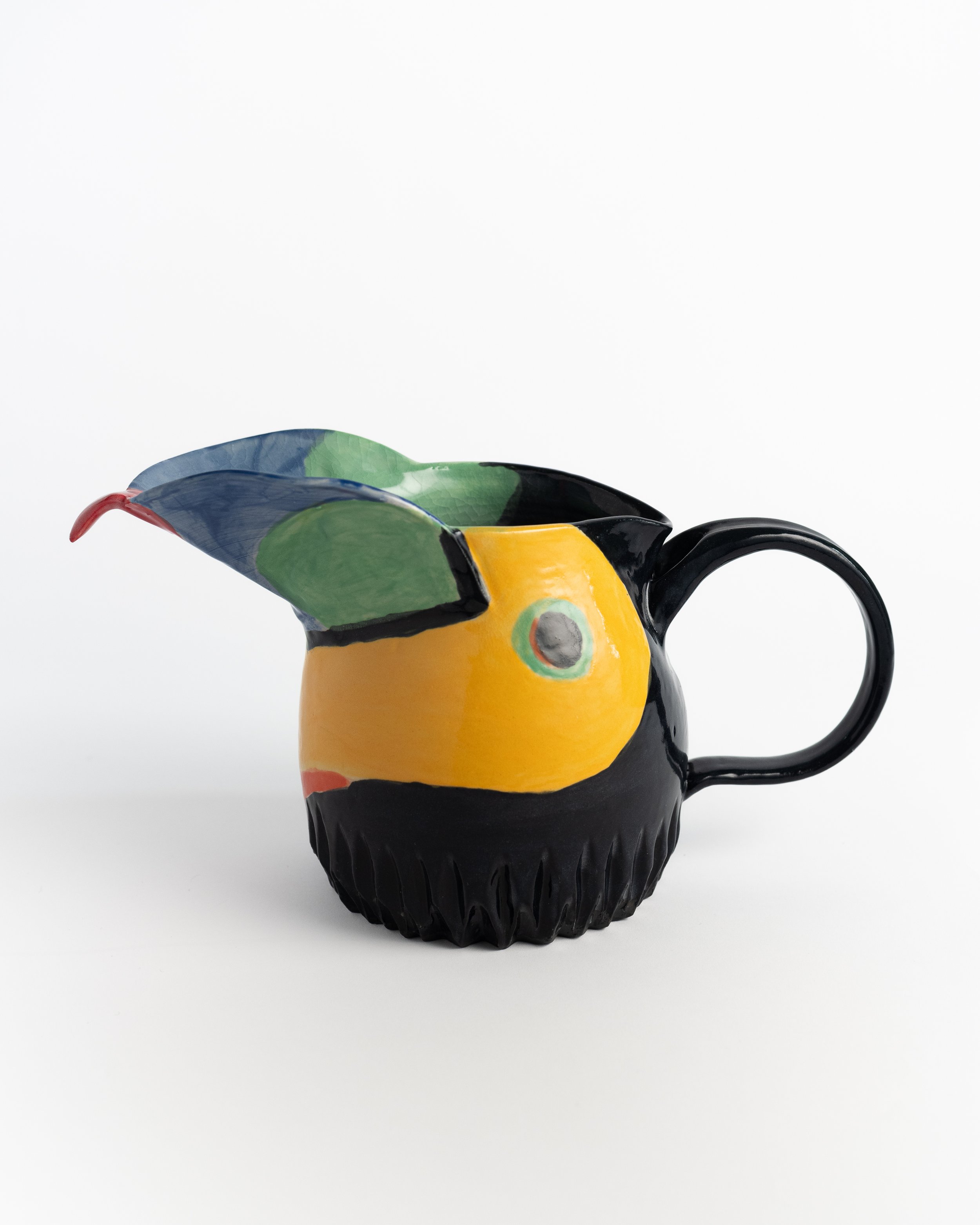 Hand-sculpted ceramic toucan pitcher in black, gold, and blue with a coral beak forming the spout.