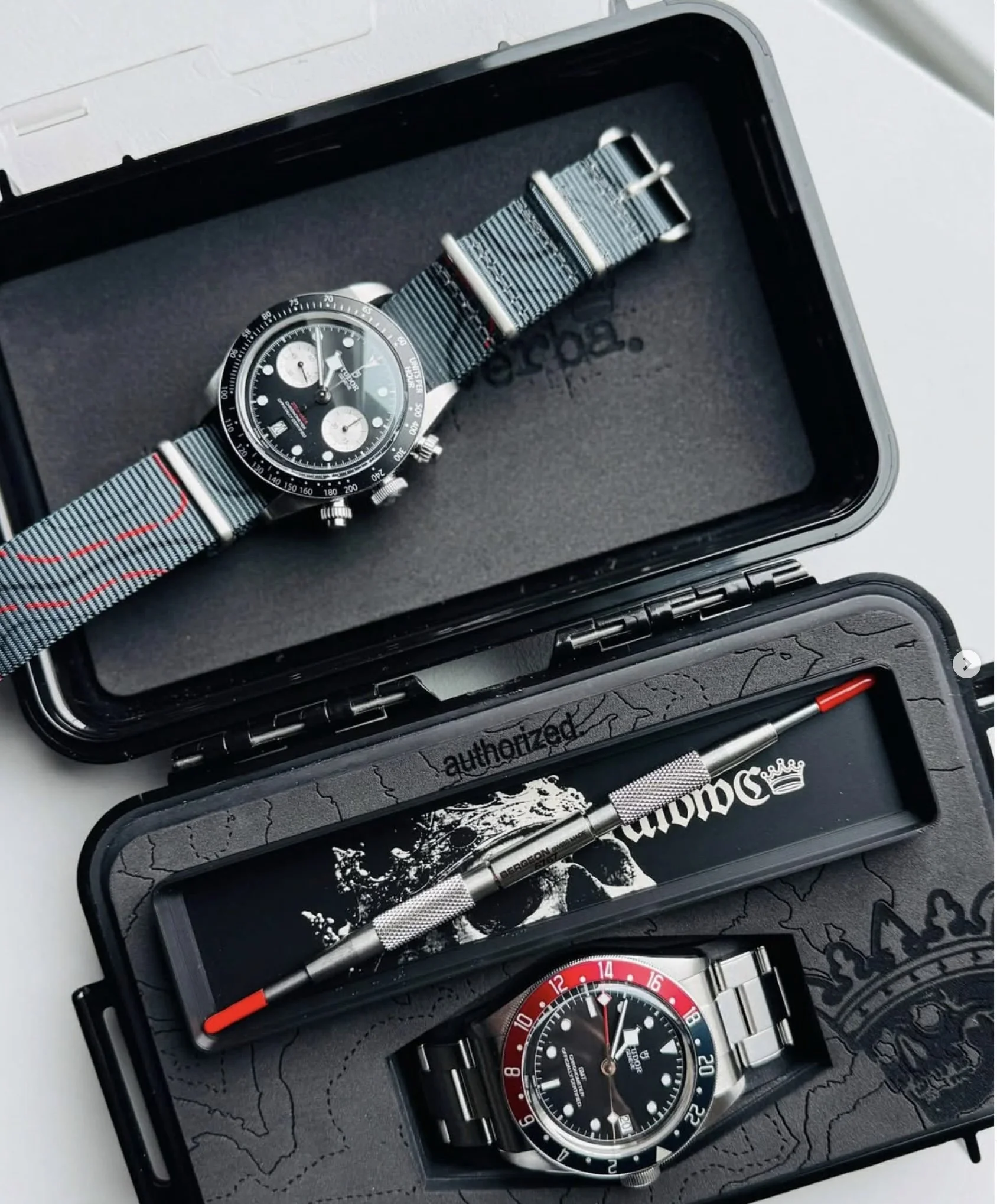 A collection of luxury watches in a black presentation case, featuring a Tudor chronograph with a striped NATO strap, a black and white themed watch with skull graphics, and a stainless steel watch with a black dial and red accents.