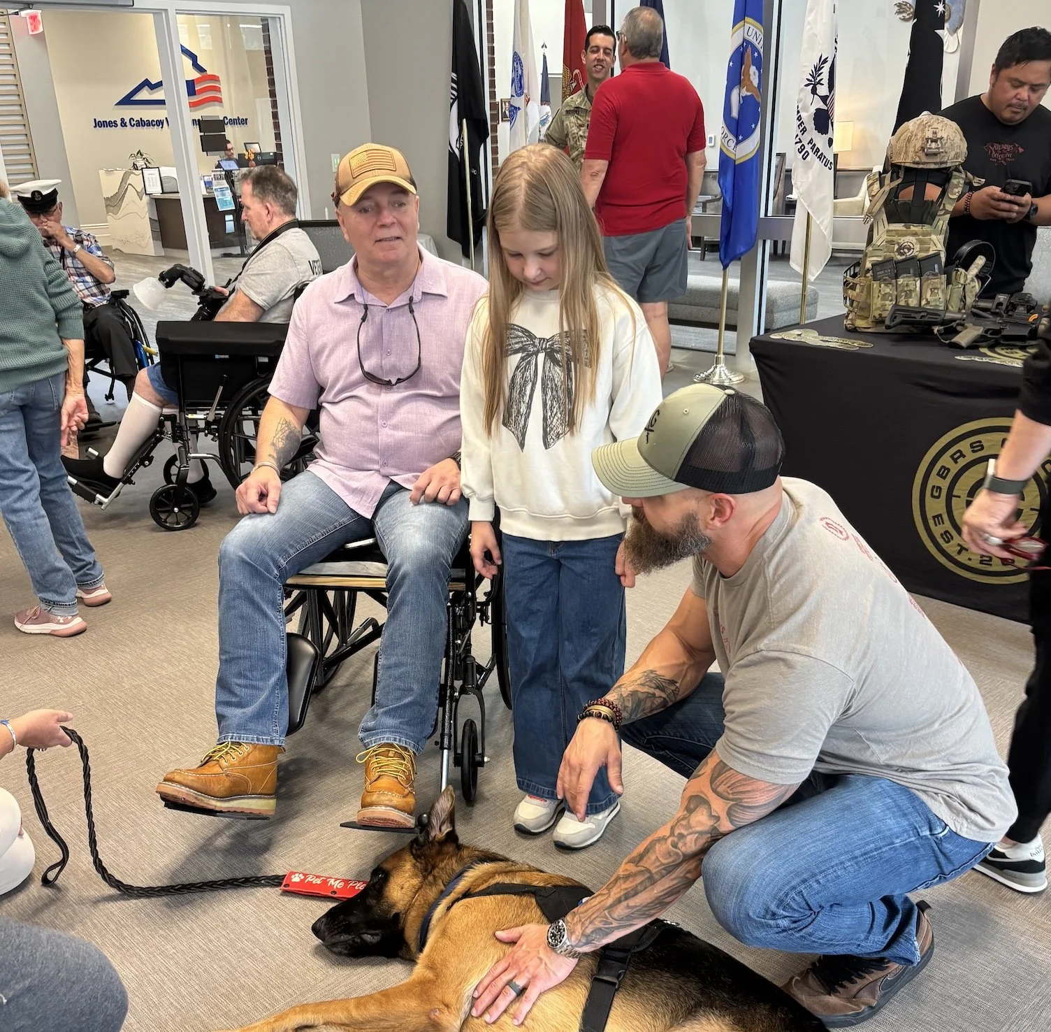 A man kneeling next to a police dog lying on the floor, with a young girl and a man in a wheelchair nearby, inside a room at the Jones & Cabcacy V.A. Center. Several people are standing and talking in the background, with flags and military displays visible.
