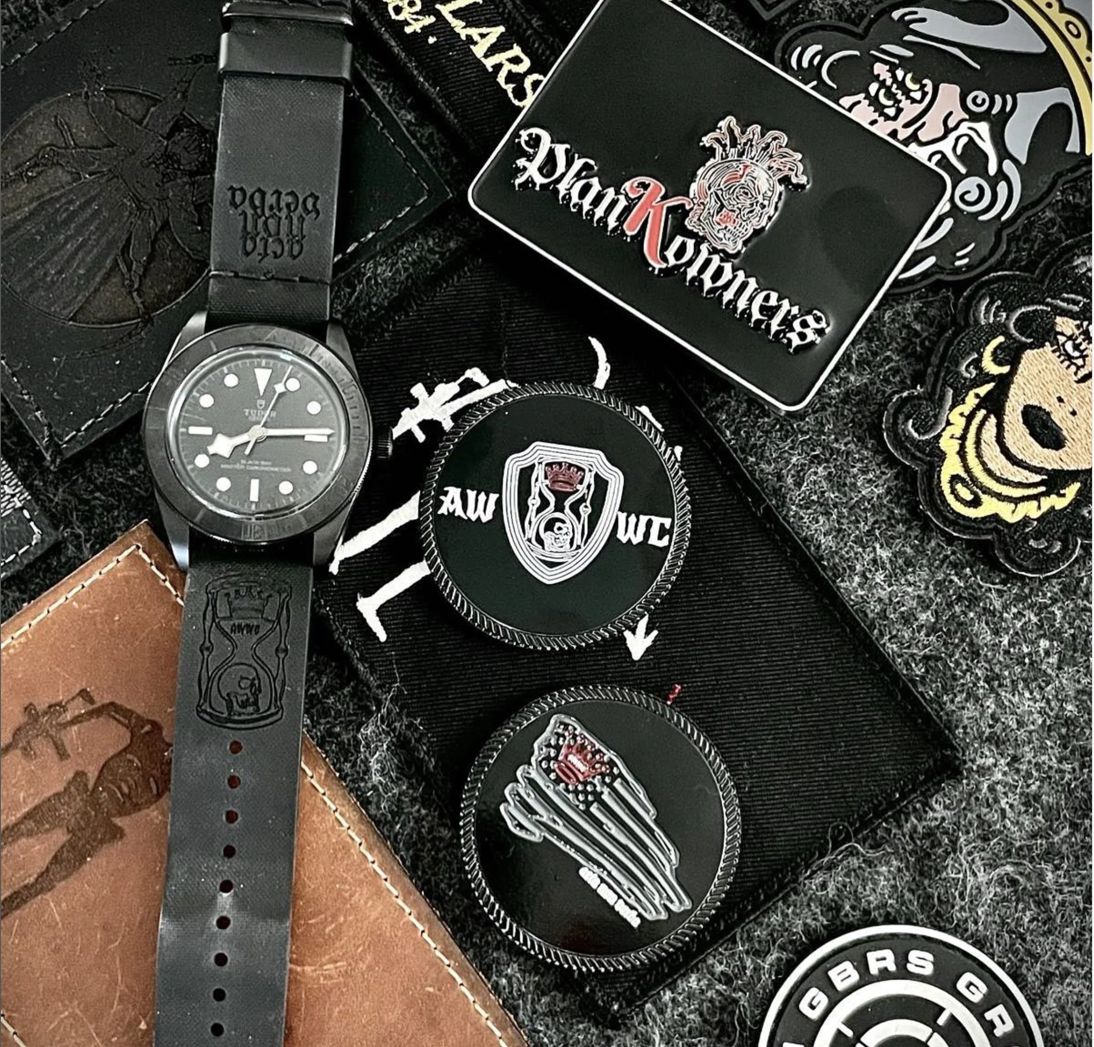 Close-up of a collection of black patches, patches with logos, watches, and a belt, arranged on a dark textured surface.
