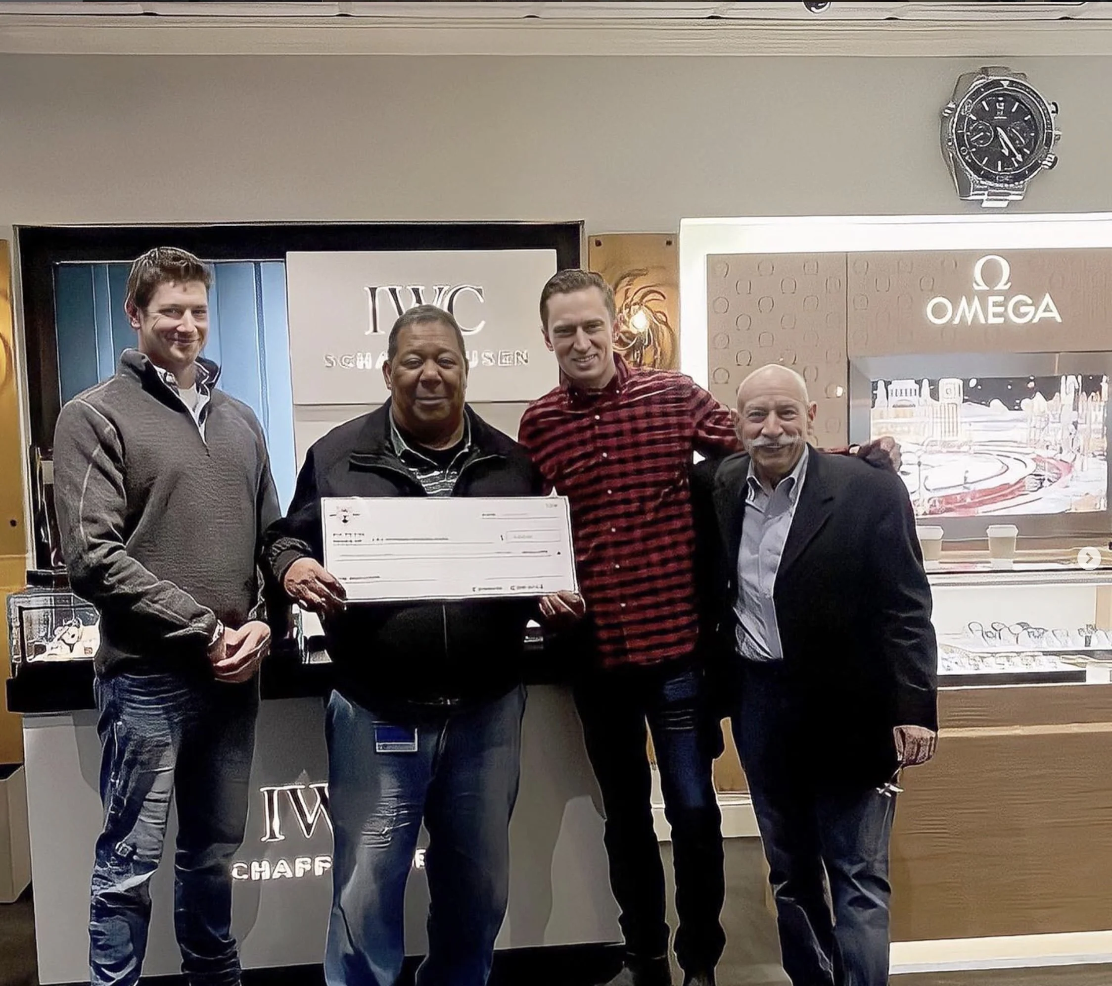 A group of five men standing inside a store, with two signs in the background that say "IWC" and "Omega." The men are smiling and holding a large check. The store display includes watches and other merchandise.