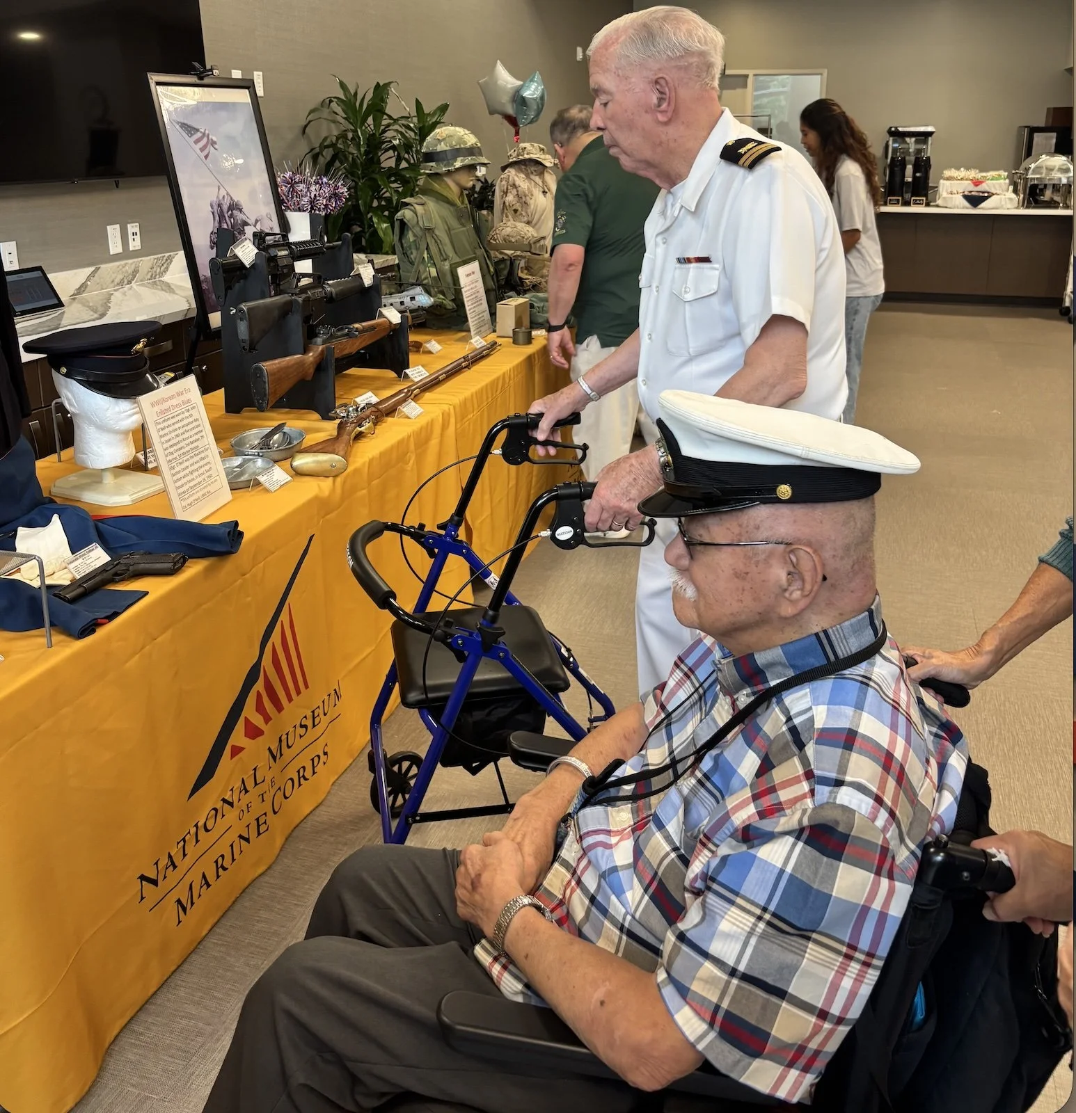 An elderly veteran in uniform wearing a white sailor's hat, sitting with a walker nearby. He is at a display table at the National Museum of the Marine Corps, looking at military artifacts. A man in the background is also inspecting items, and there are balloons and other people in the room.