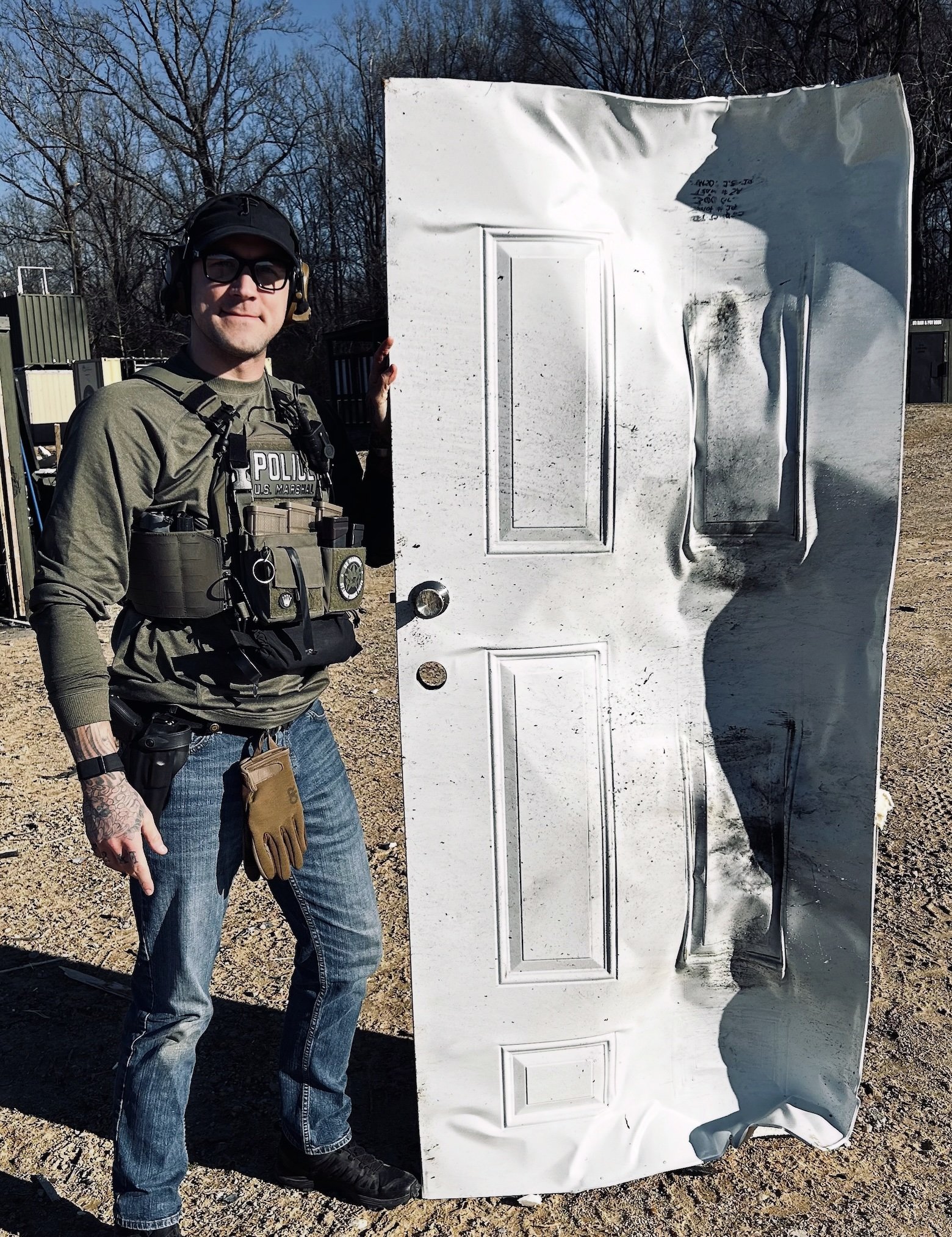 A police officer in tactical gear standing next to a damaged white door outdoors on a sunny day.