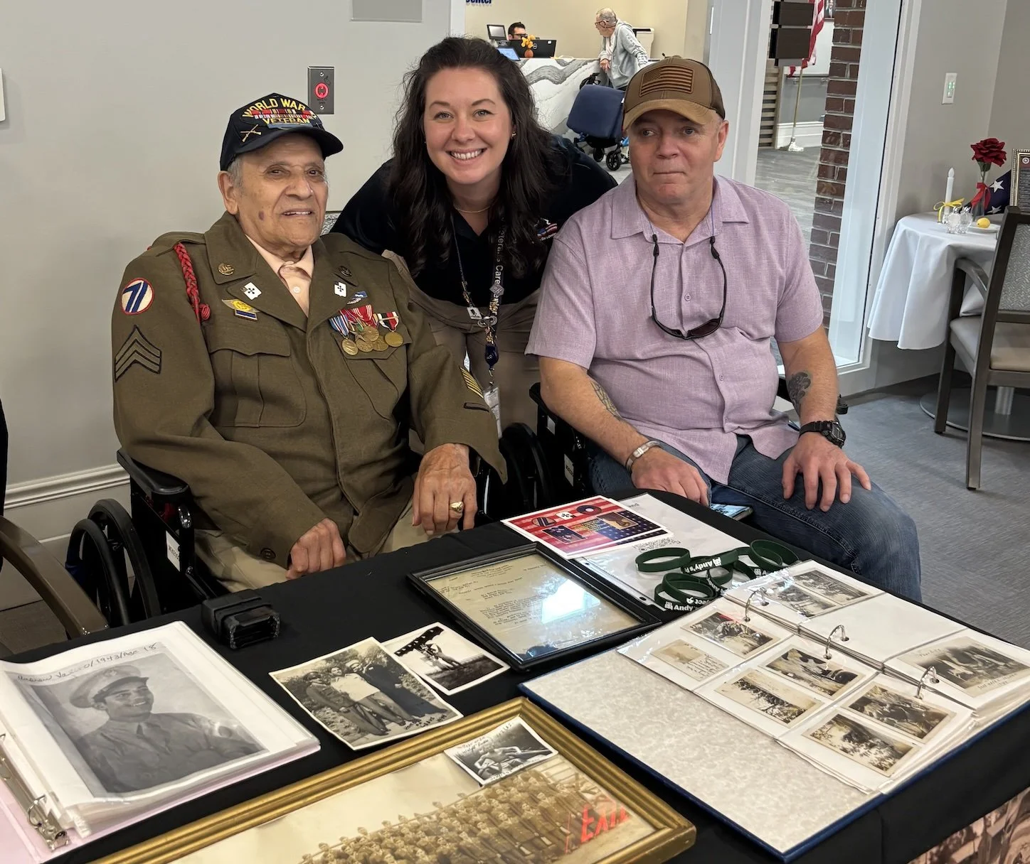 A smiling woman is standing between an elderly man in a military uniform and a middle-aged man wearing a light purple shirt. The elderly man is in a wheelchair, and there are photographs and memorabilia on the table in front of them suggesting a reunion or commemorative event.
