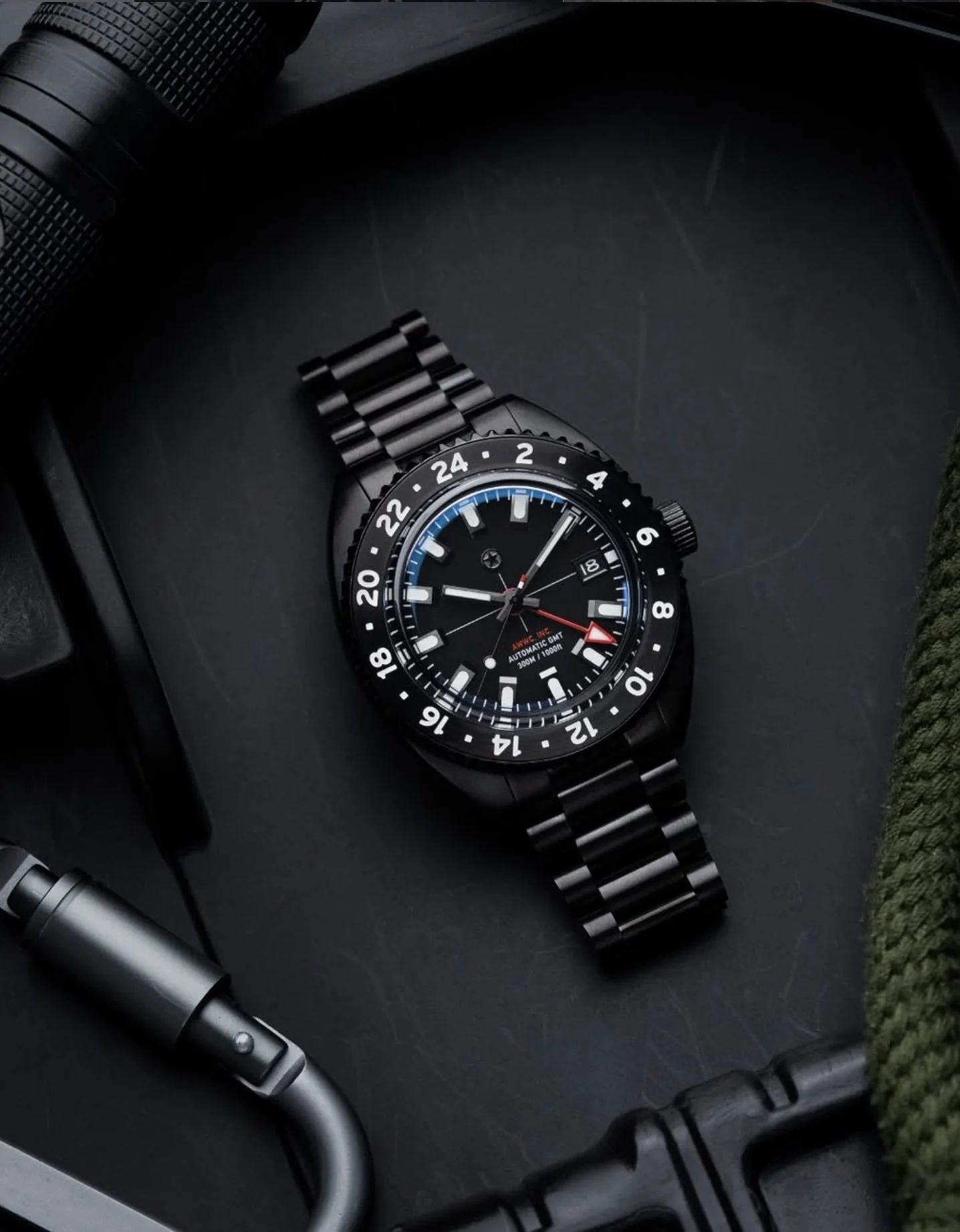 Black wristwatch with a metal band, multiple dials, and a 24-hour bezel, lying on a dark surface surrounded by a flashlight, a flashlight clip, and a piece of green fabric.