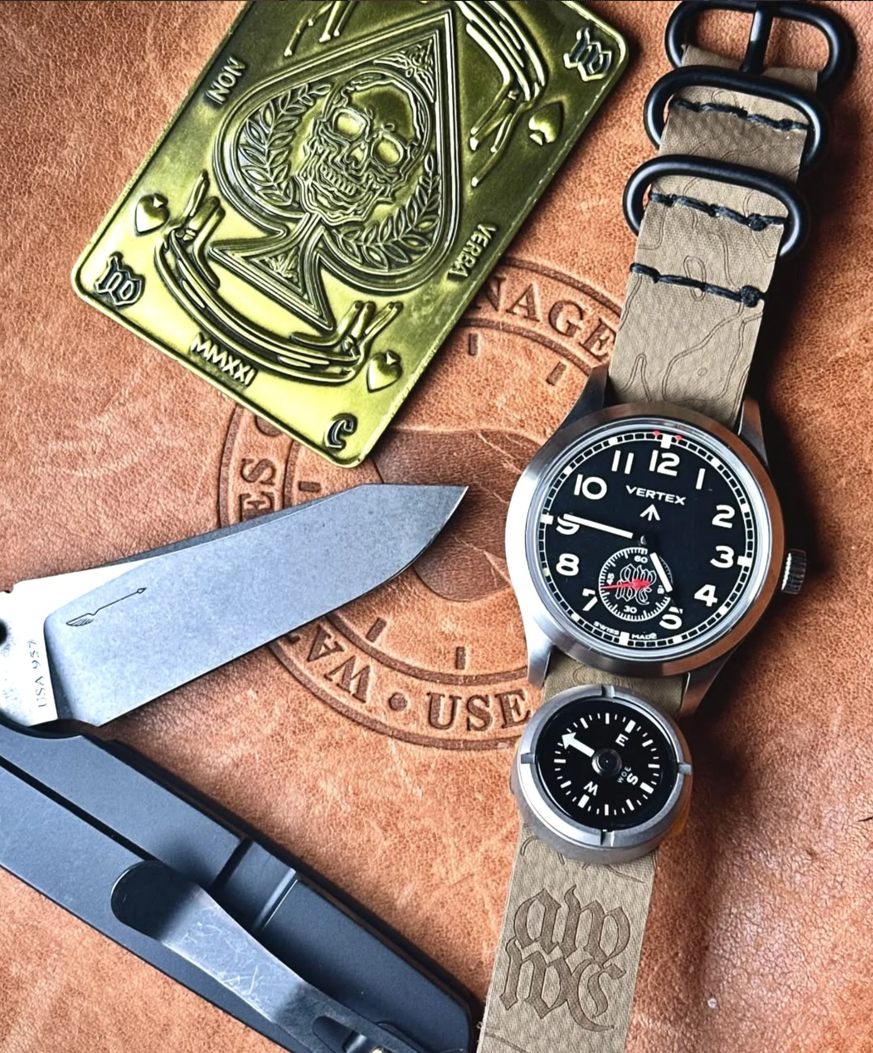 Close-up of a wristwatch with a black dial and a terrain-patterned band, a metal knife with a black handle, a gold-colored playing card featuring a skull design, and a compass, all placed on a leather surface with engraved text.