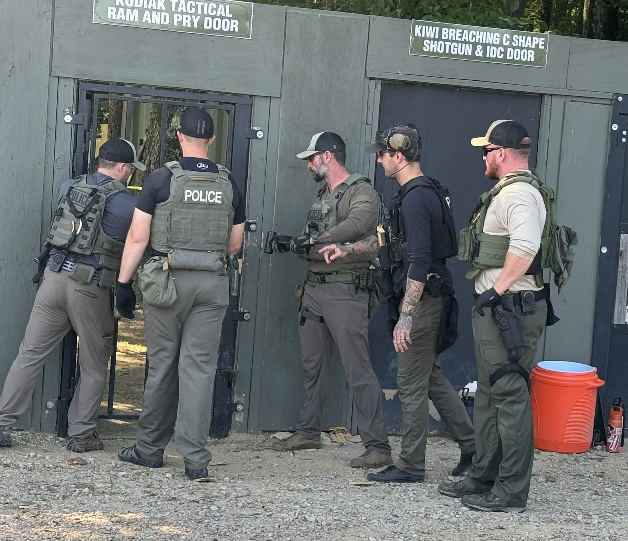 Five tactical officers in uniform with guns and gear, standing outside a building labeled with signs regarding tactical doors and shotgun doors; they appear to be preparing or discussing a tactical operation.