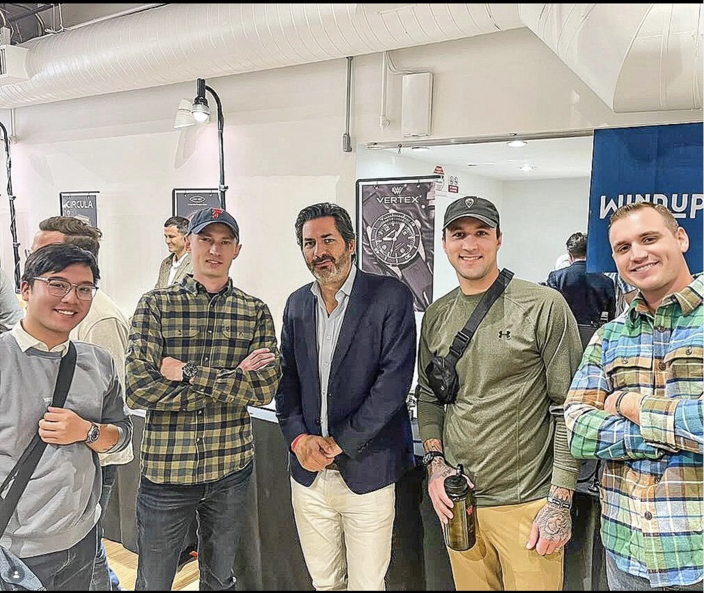 Group of men gathered together at an indoor event, some smiling and looking at the camera, with banners and equipment visible in the background.