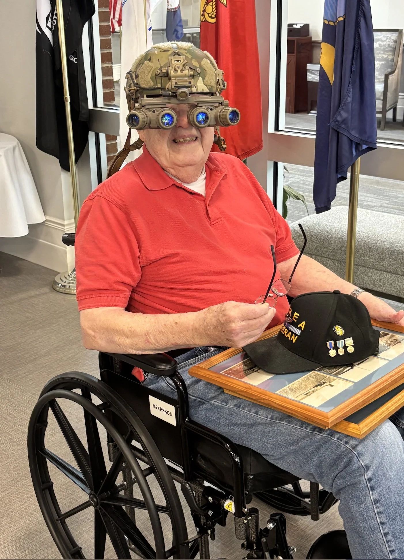 An elderly man sitting in a wheelchair, wearing a camouflage helmet with night-vision goggles, a red polo shirt, and holding glasses. On the table in front of him is a black military cap with medals and pins. The background features flags and a window.