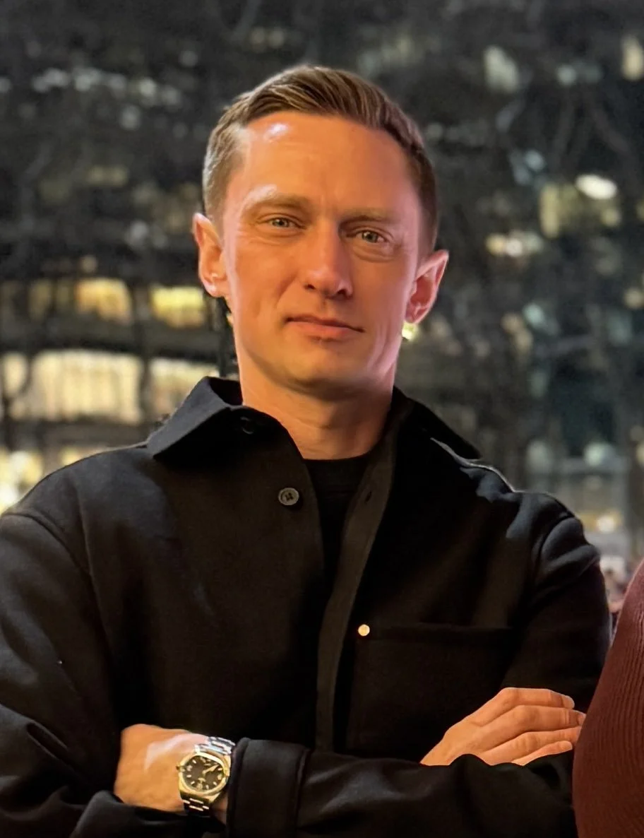 A man with short, light brown hair and fair skin, wearing a black shirt, standing with arms crossed outdoors at night.