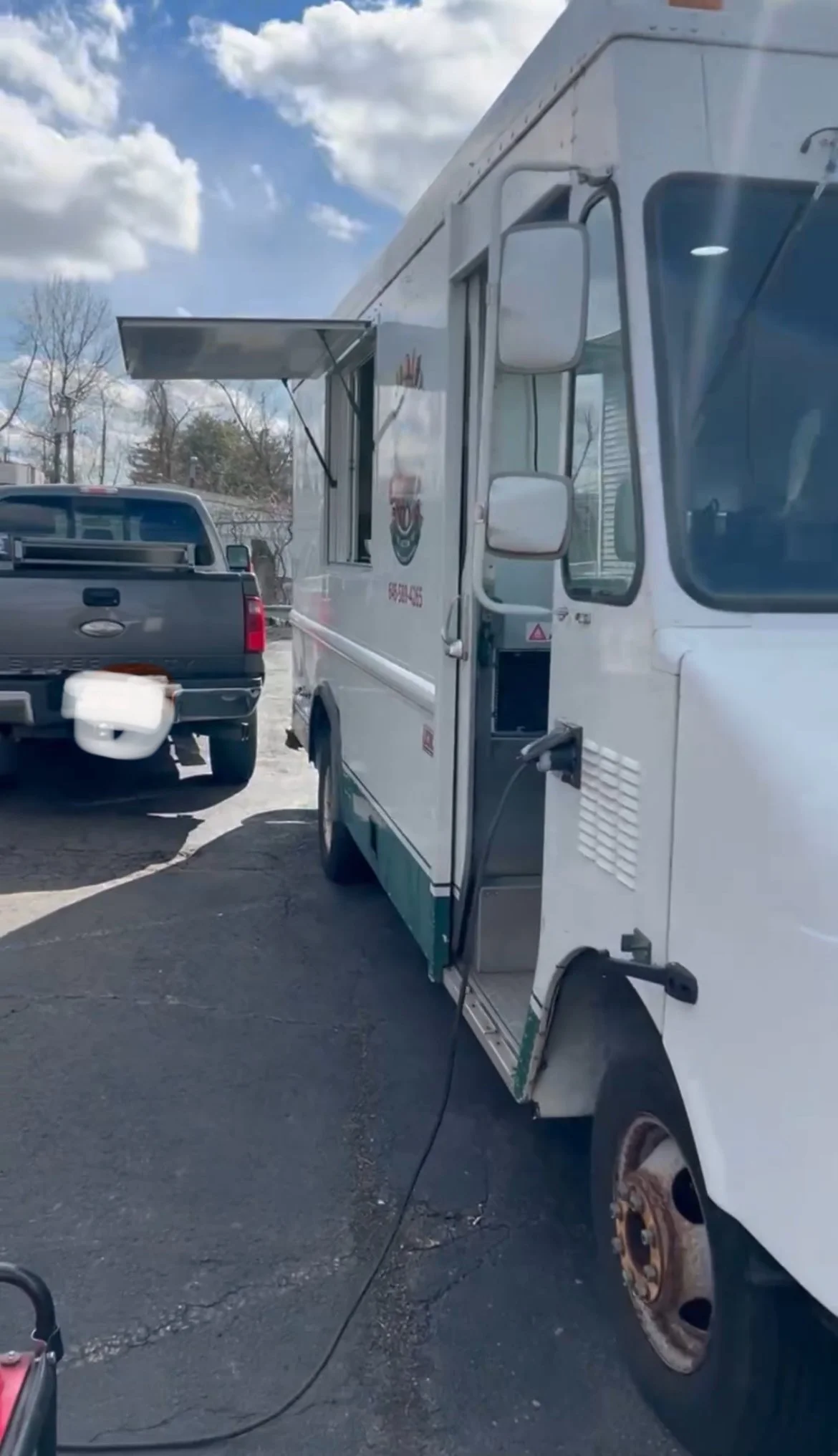 A white food truck with an open serving window, parked in a lot with a pickup truck nearby.