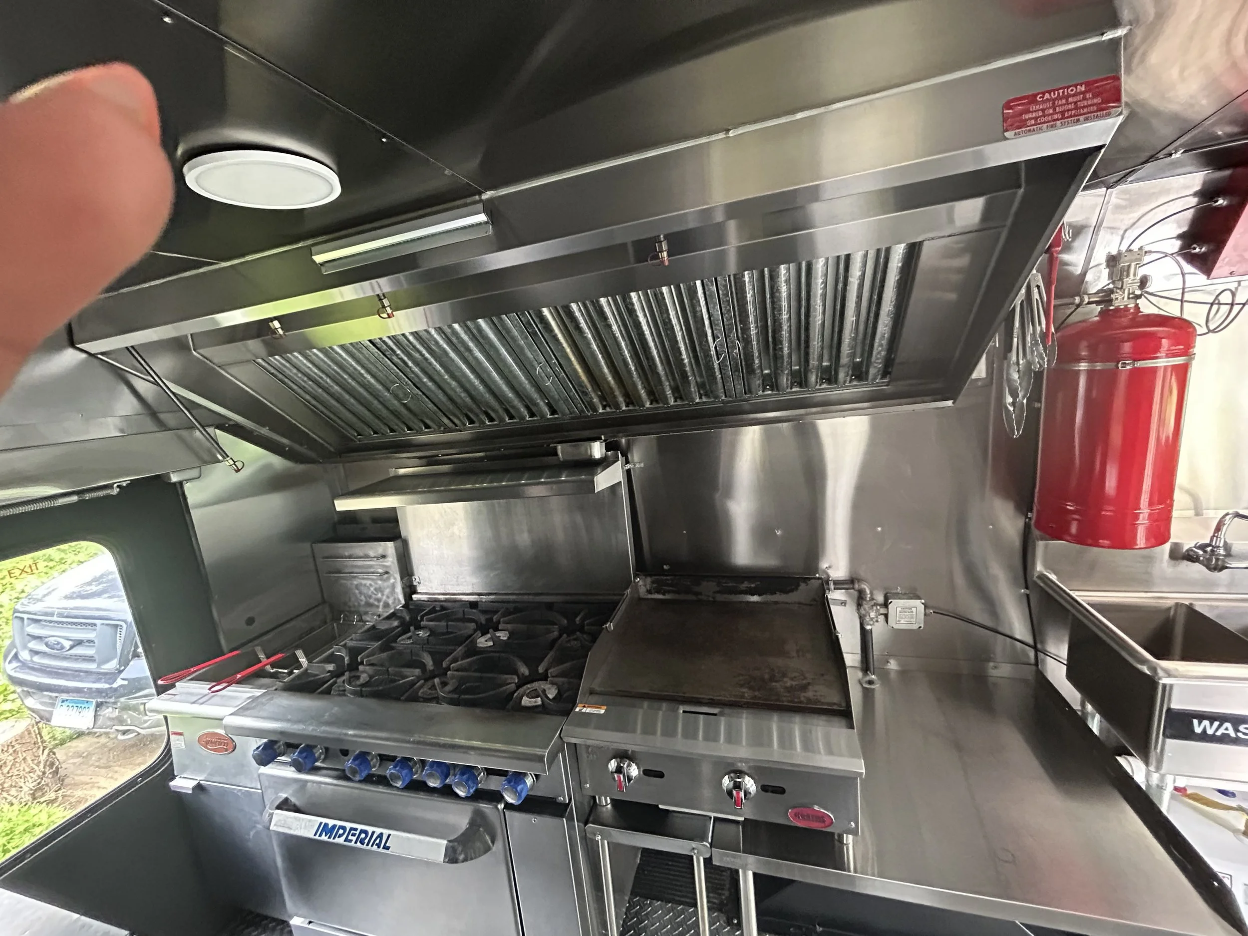 Commercial kitchen with stainless steel appliances, six-burner stove, flat grill, exhaust hood, red fire extinguisher, and nearby sink.