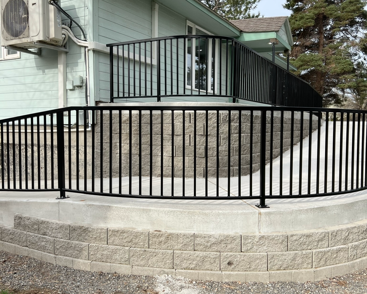 Black metal railing installed around the exterior of a house, on a raised concrete porch with a stone foundation, with snow on the ground.
