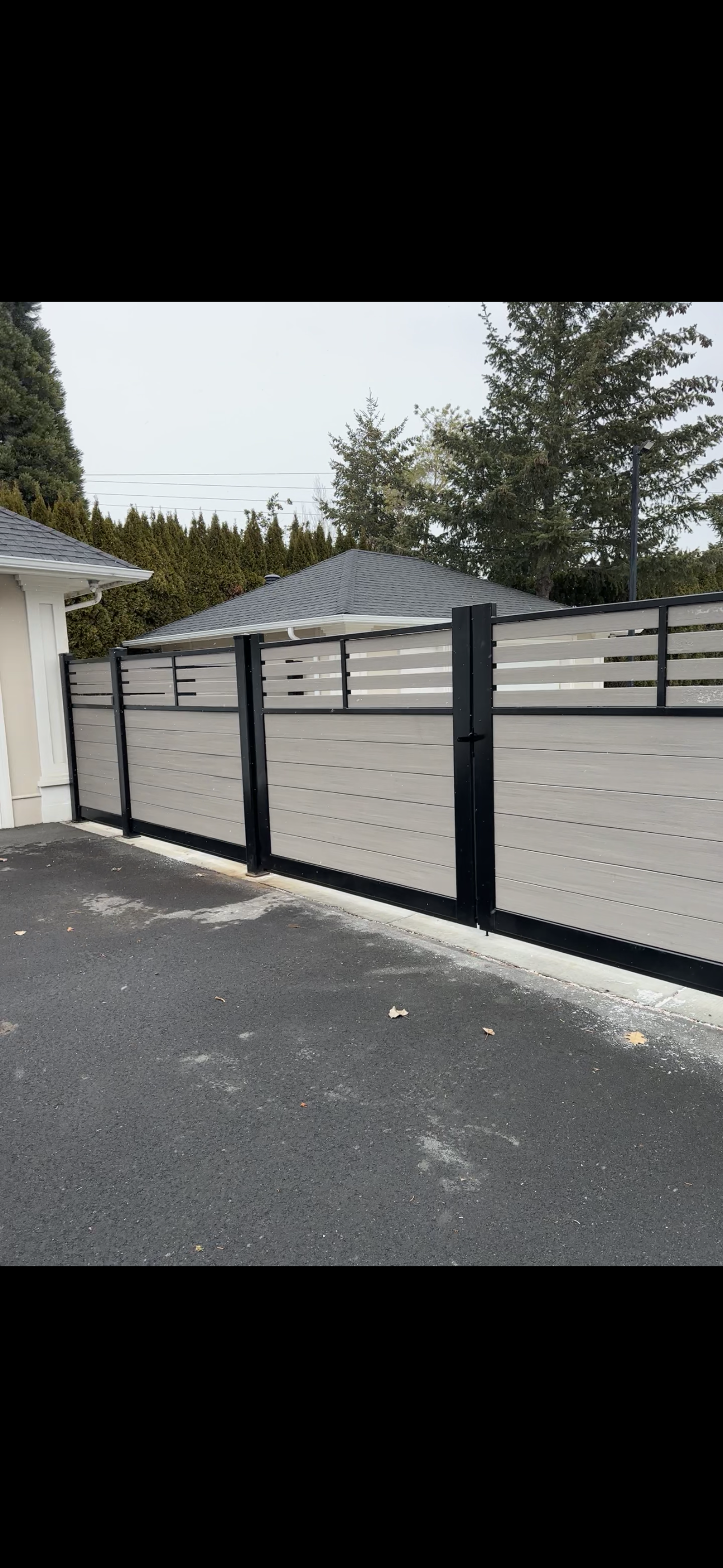 A beige and black metal fence with horizontal slats in a residential area, adjacent to a paved driveway and a house with a gray roof, with trees in the background.