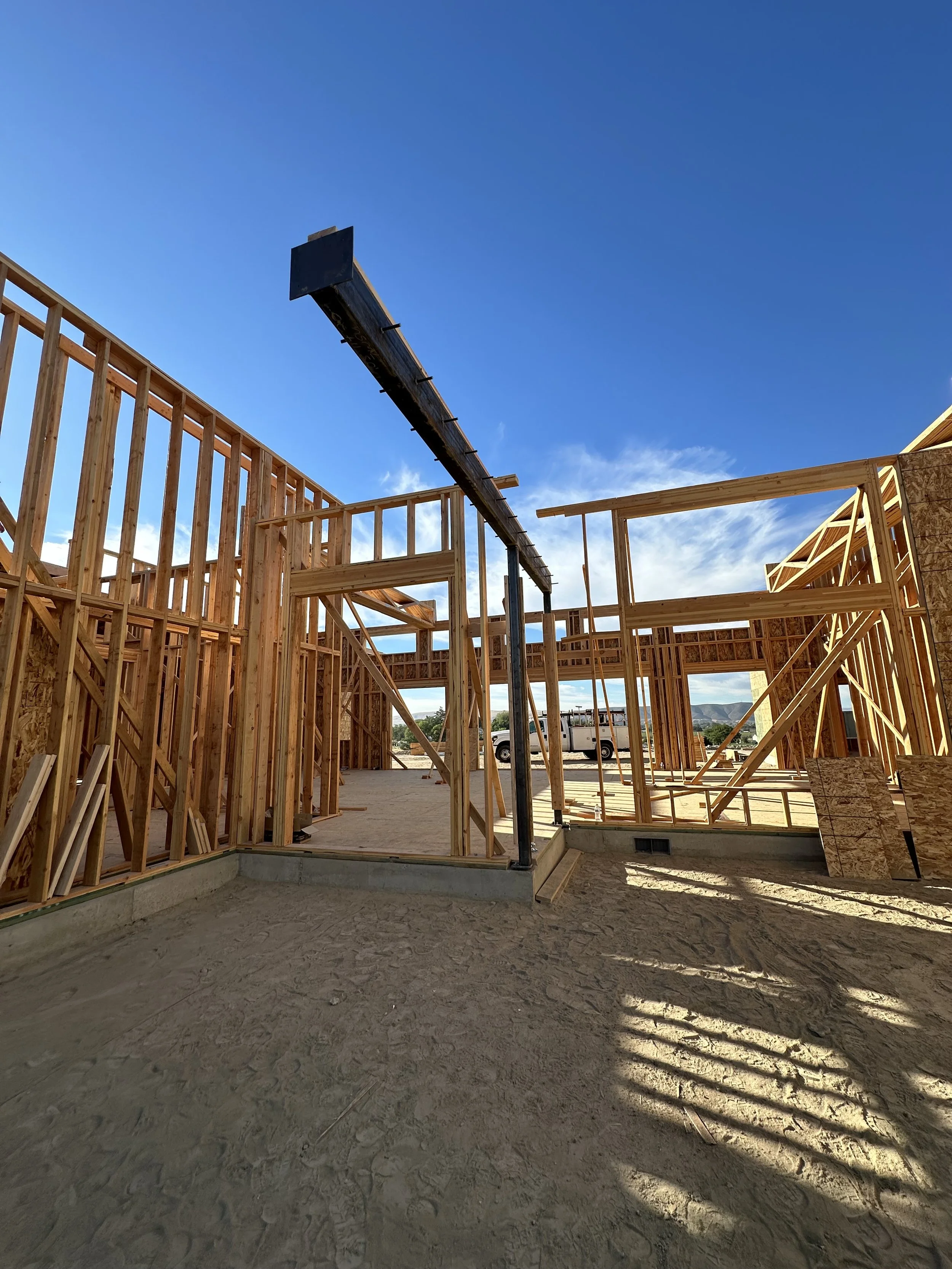 Wood-frame construction site with partially built walls and a wooden roof structure, under a blue sky with some clouds.