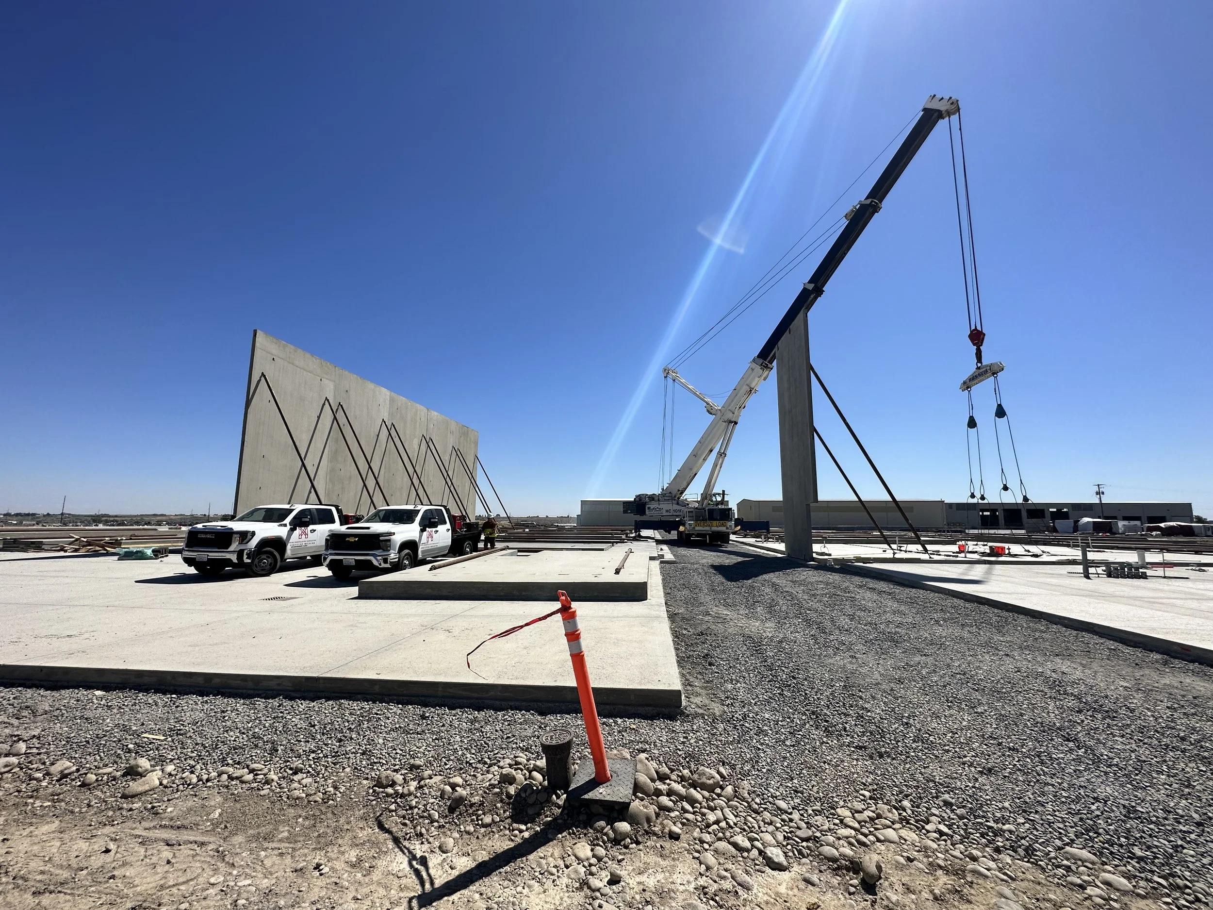 Construction site with large crane lifting materials, parked pickup trucks, and a partially constructed wall with diagonal supports under a clear blue sky.