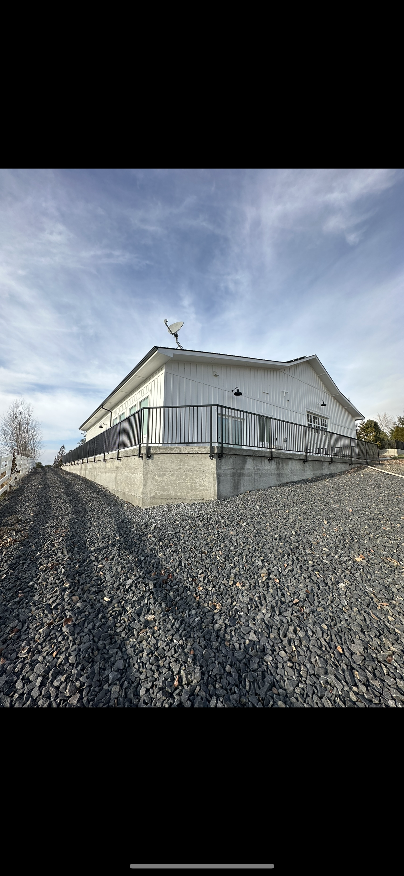 A modern house with white siding and a sliding glass door, built on a concrete foundation with a metal railing, situated on a gravel lot under a partly cloudy sky.