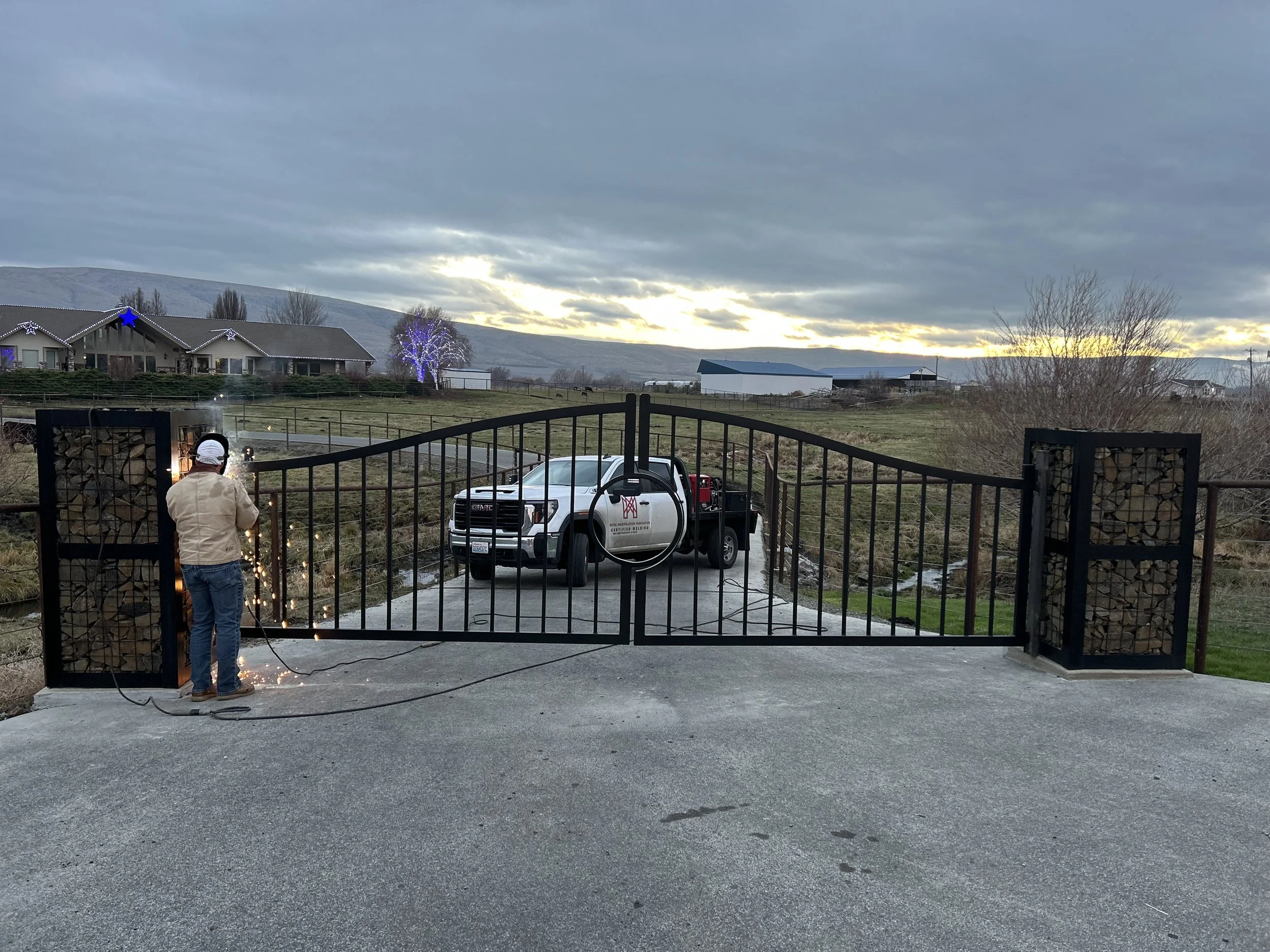 A person welding on a gate with sparks flying, a pickup truck parked behind on a driveway, and houses with Christmas lights in the background at sunset.