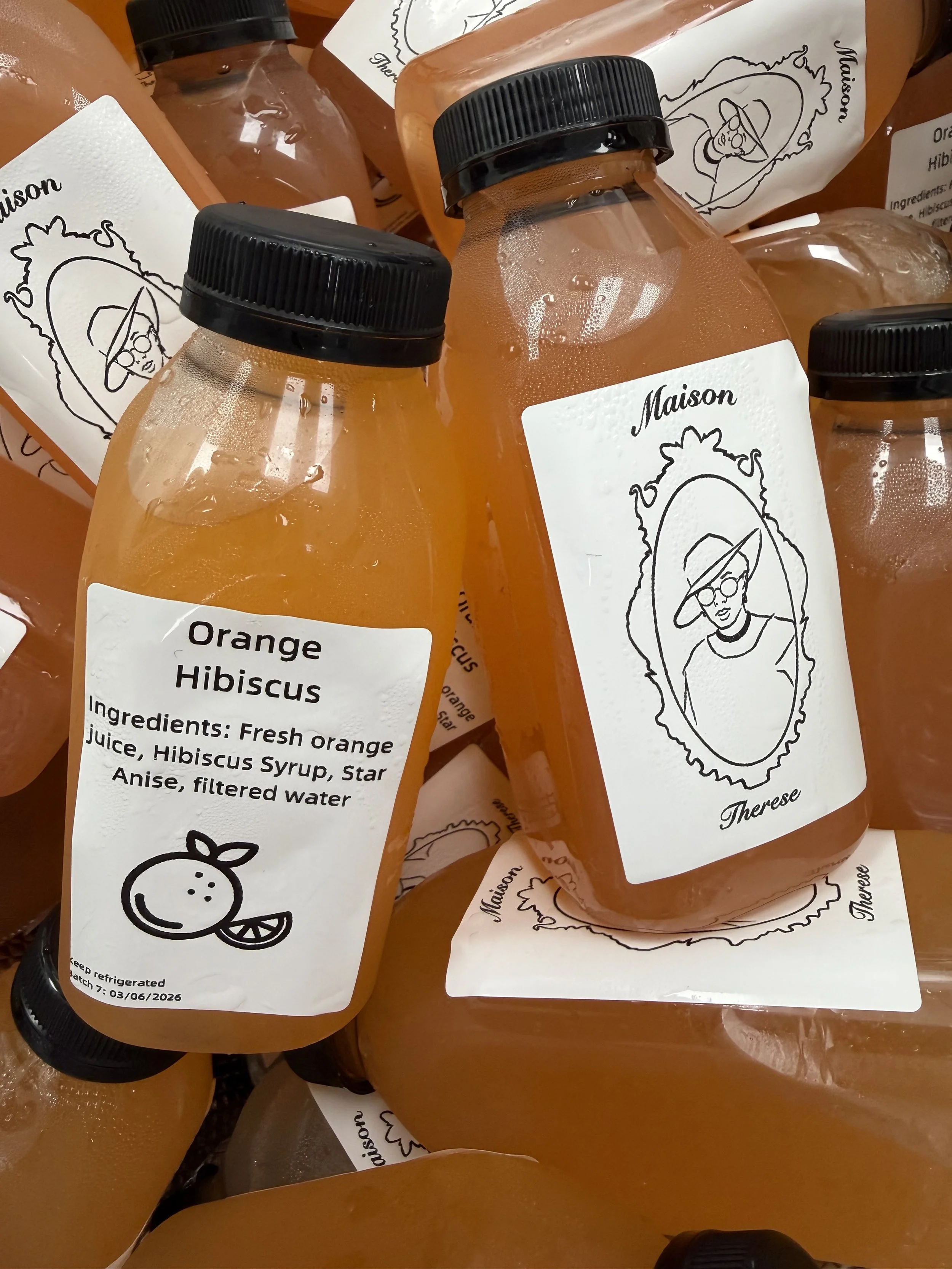 Several bottles of orange hibiscus juice with black caps, some with labels showing a line drawing of a woman in a hat and glasses, and ingredients listed as fresh orange juice, hibiscus syrup, star anise, and filtered water.