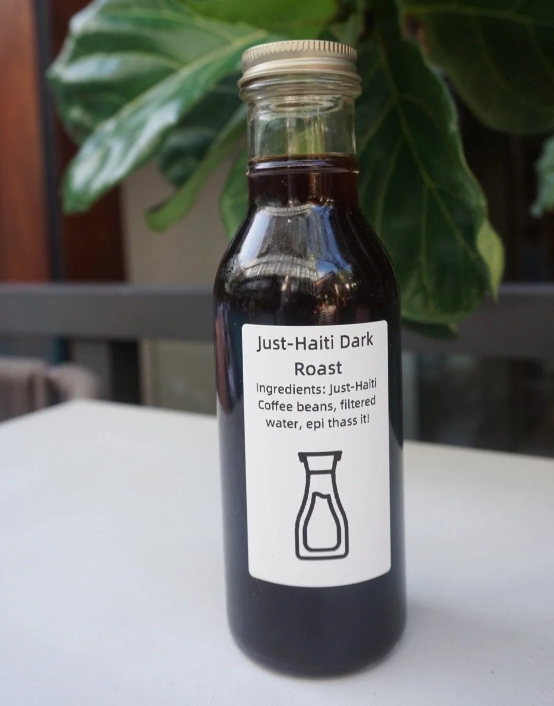 A glass bottle labeled "Just-Haiti Dark Roast" containing dark liquid, with a white label and a simple icon of a coffee pot, set against a background of green leaves.