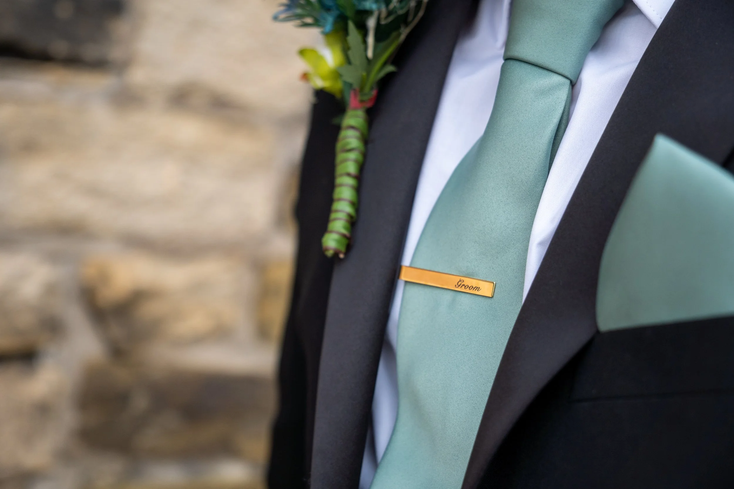 Close-up of groom's attire with a light green tie, gold 'Groom' name tag, and boutonniere, against a textured stone wall background.