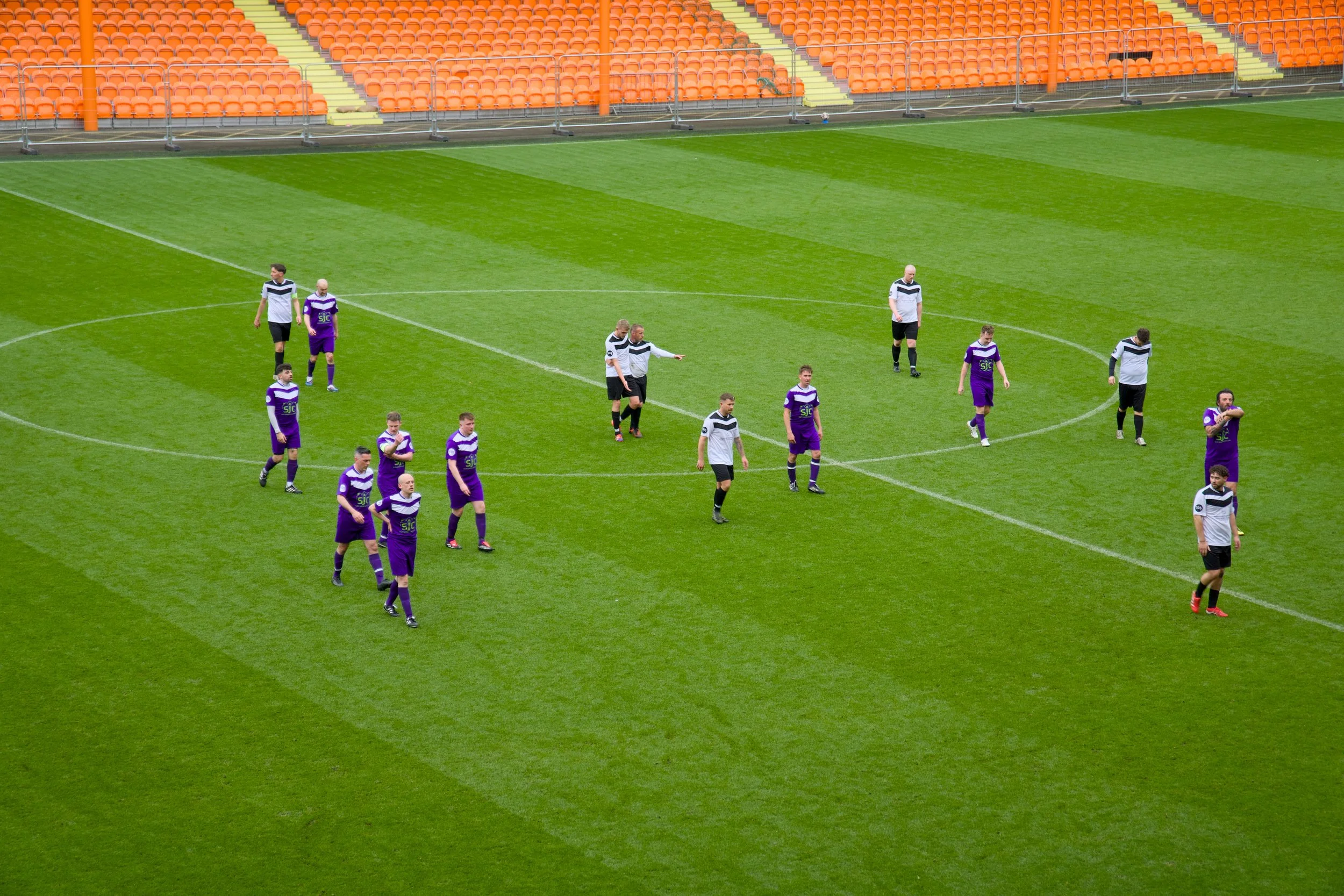 A soccer team on a green field with orange stadium seats in the background. Some players are wearing purple jerseys and some in black and white jerseys, walking or standing on the field.