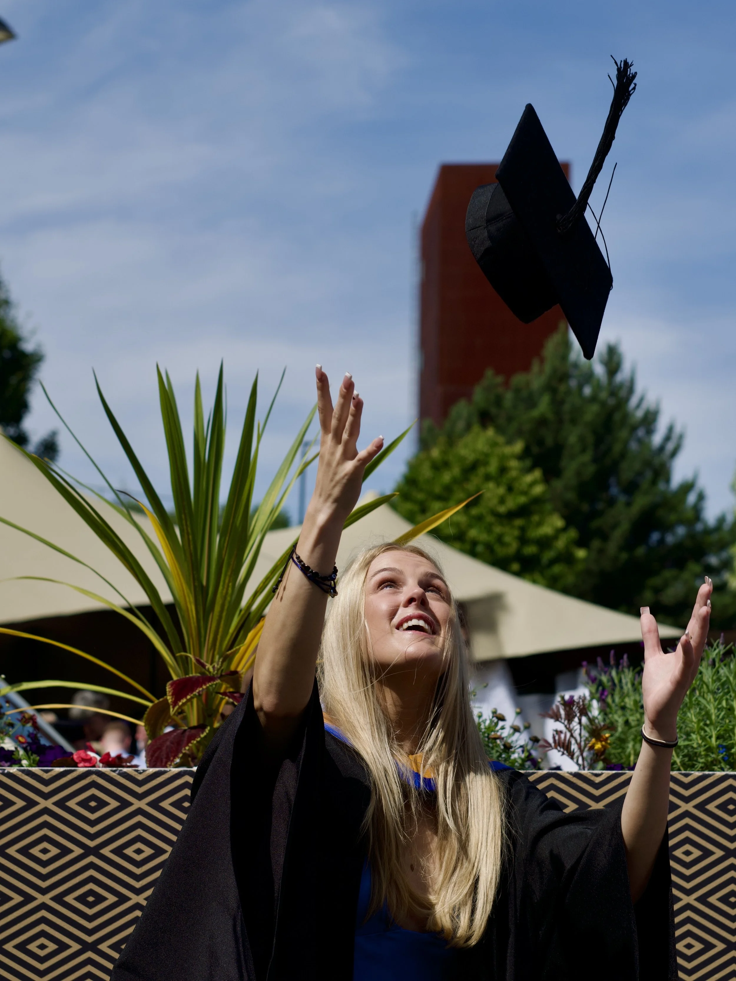 A young woman with long blonde hair celebrating her graduation, wearing a cap and gown, throwing her cap into the air outdoors during daytime with a clear blue sky and trees in the background.