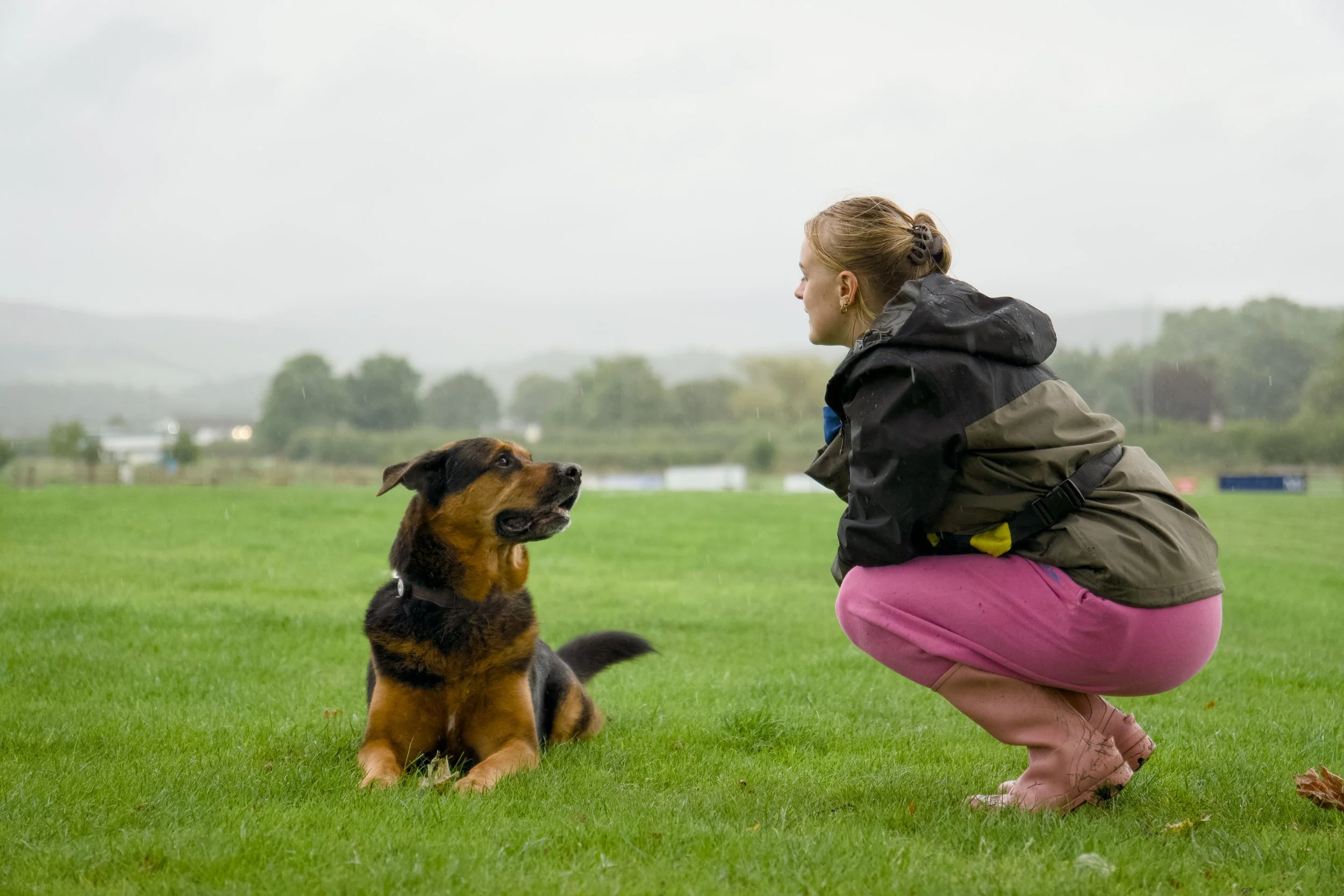 Woman in outdoor rain gear kneeling on grass, looking at dog sitting in front. Overcast sky with background of trees and field.